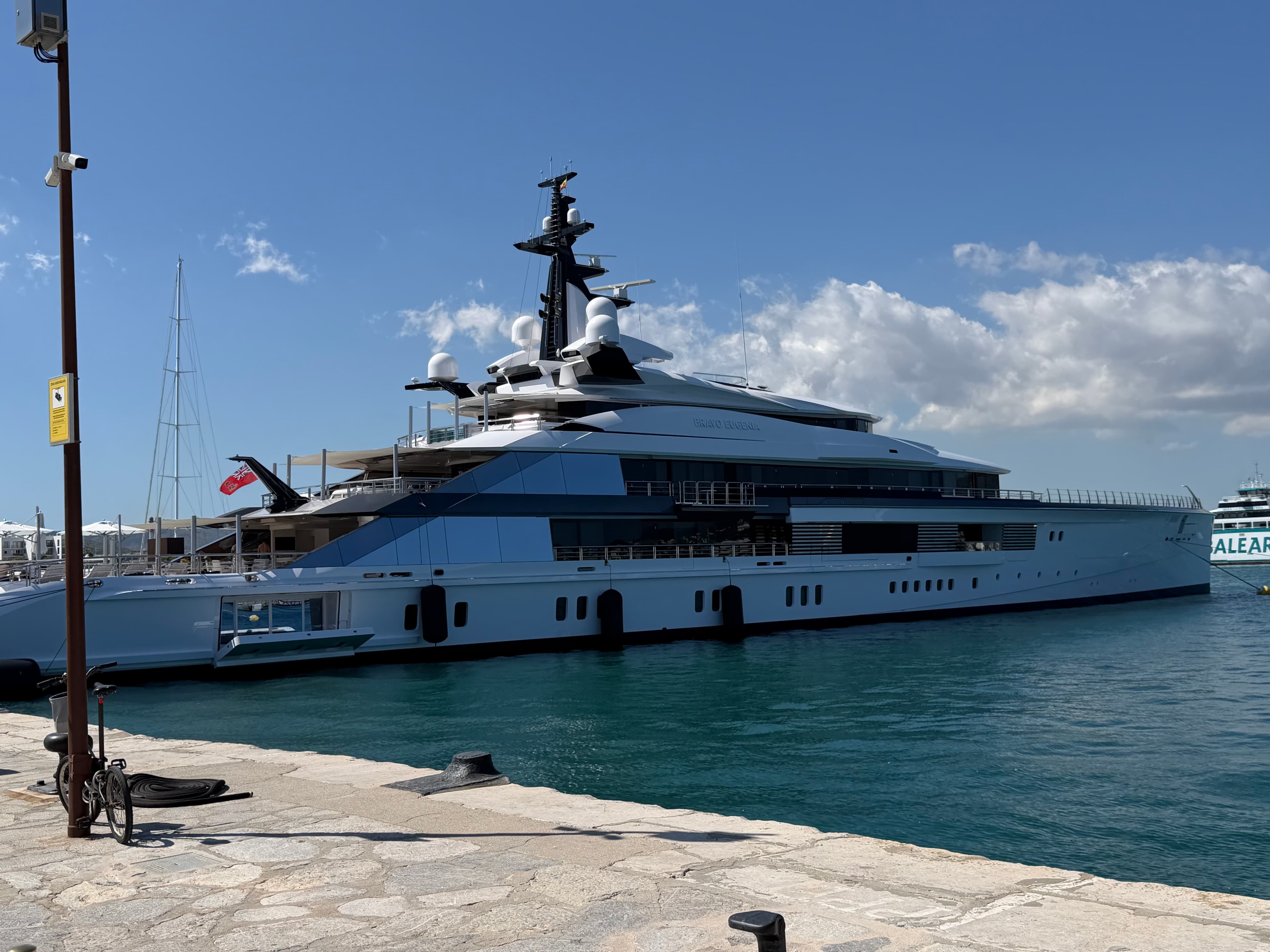 Massive modern superyacht with dark navy and white hull docked at a sunny Mediterranean marina under a blue sky with scattere
