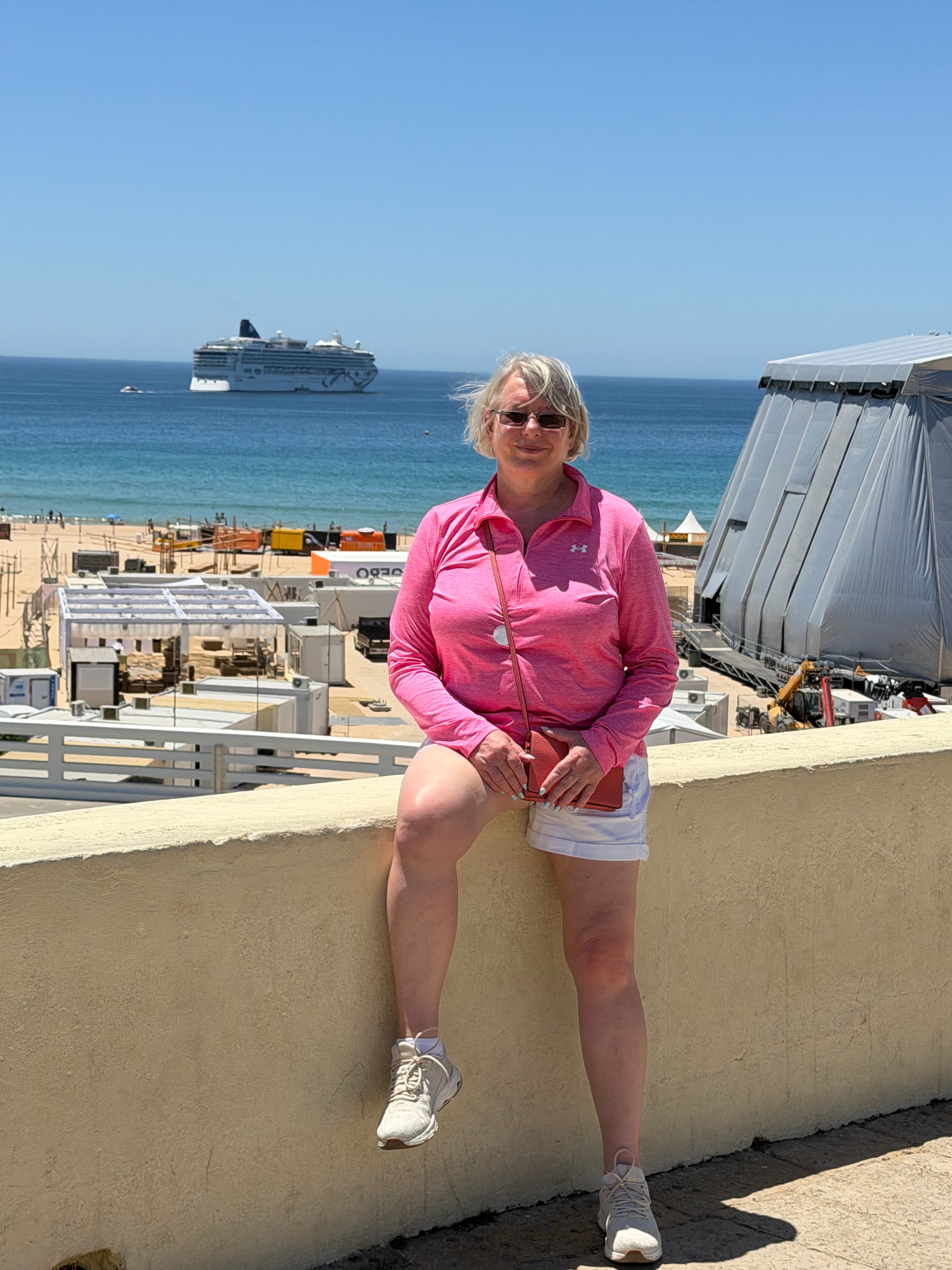 Woman in pink athletic top leaning on a sun-bleached wall overlooking a sandy beach, event structures, and a cruise ship on v