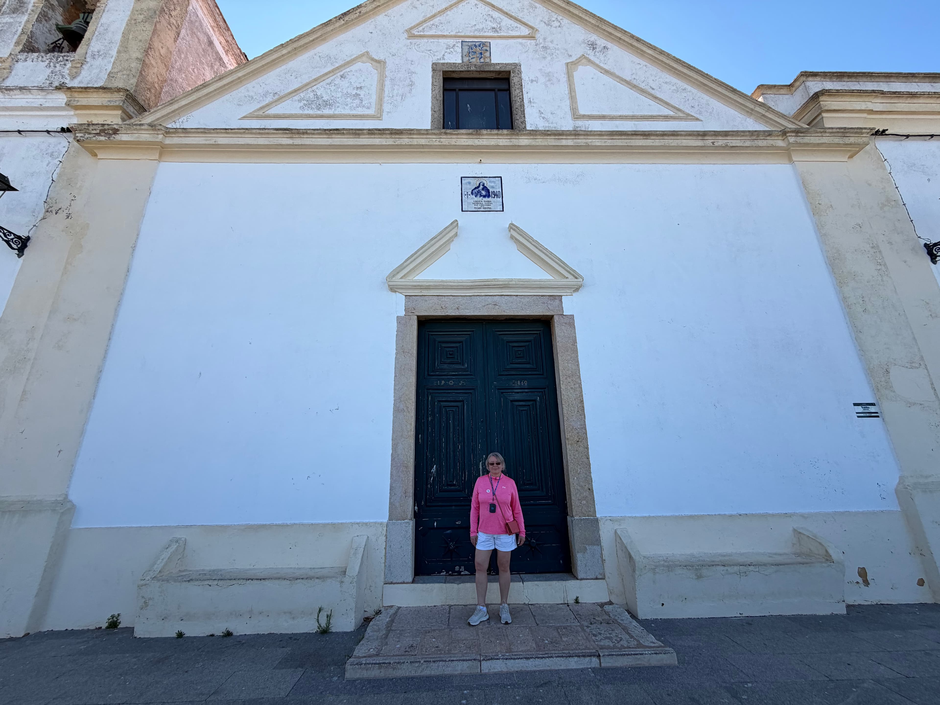 Woman in pink top standing before a whitewashed Portuguese chapel with dark green wooden doors and stone pediment detailing,