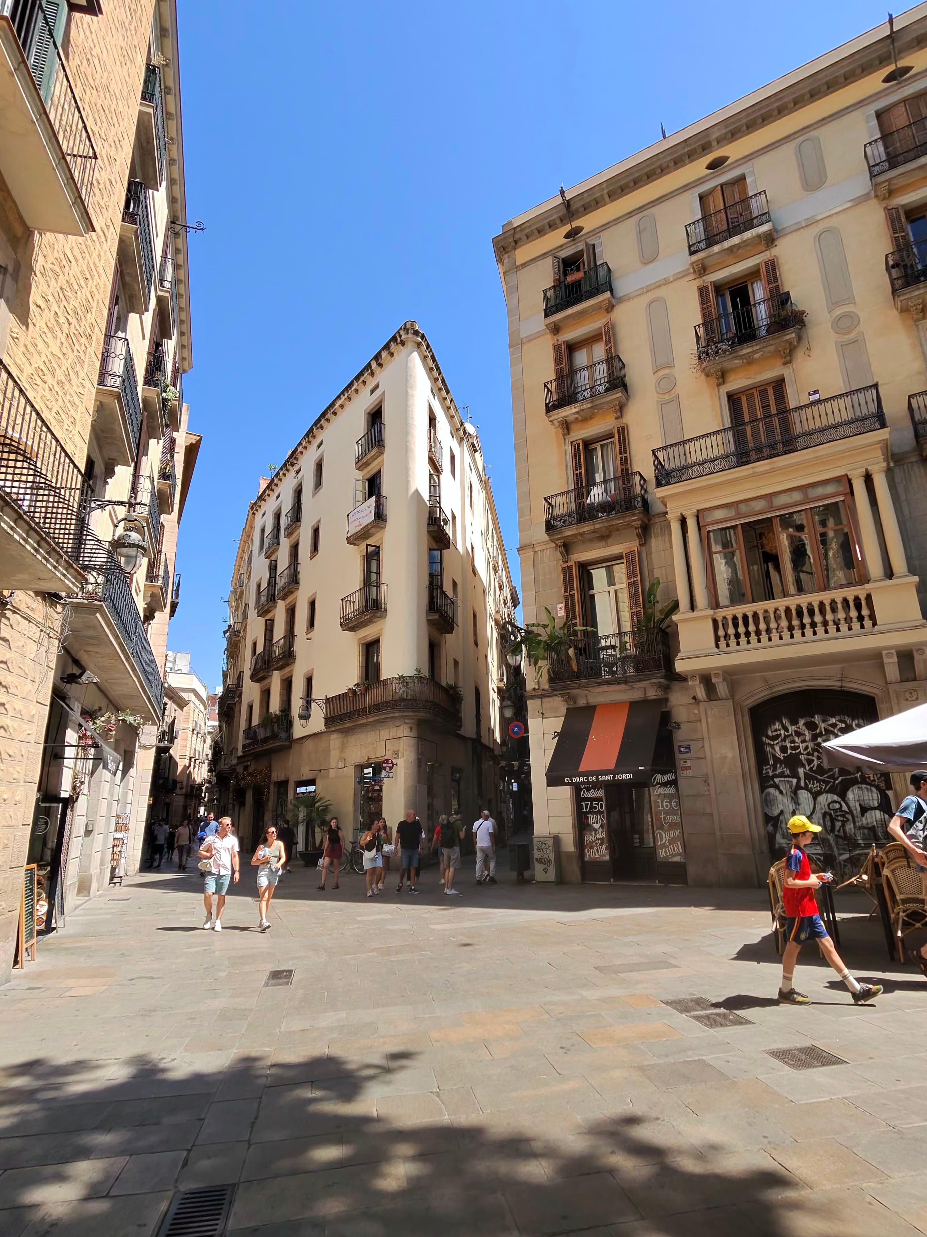Sunny pedestrian plaza in Barcelona's Gothic Quarter with ornate balconied buildings converging at a narrow wedge-shaped corn