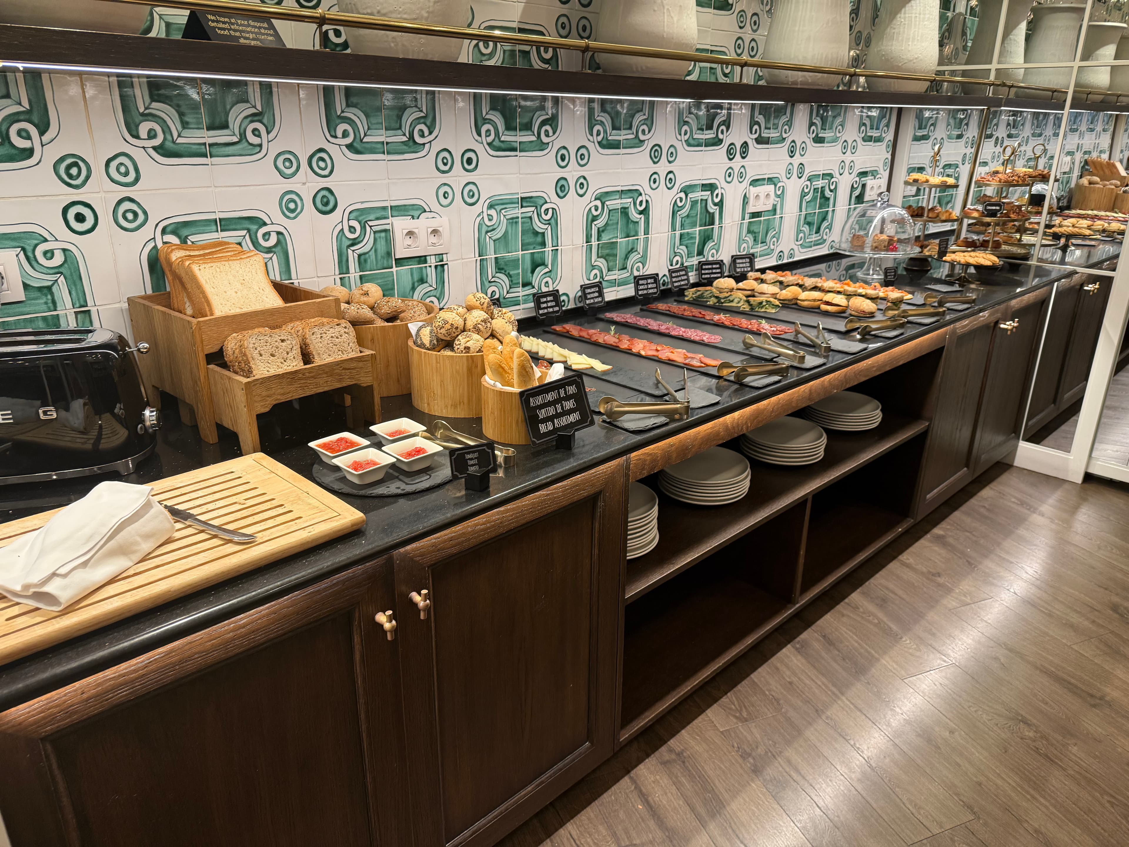 Hotel breakfast buffet spread with artisan breads, smoked salmon, pastries, and condiments against bold green-patterned decor