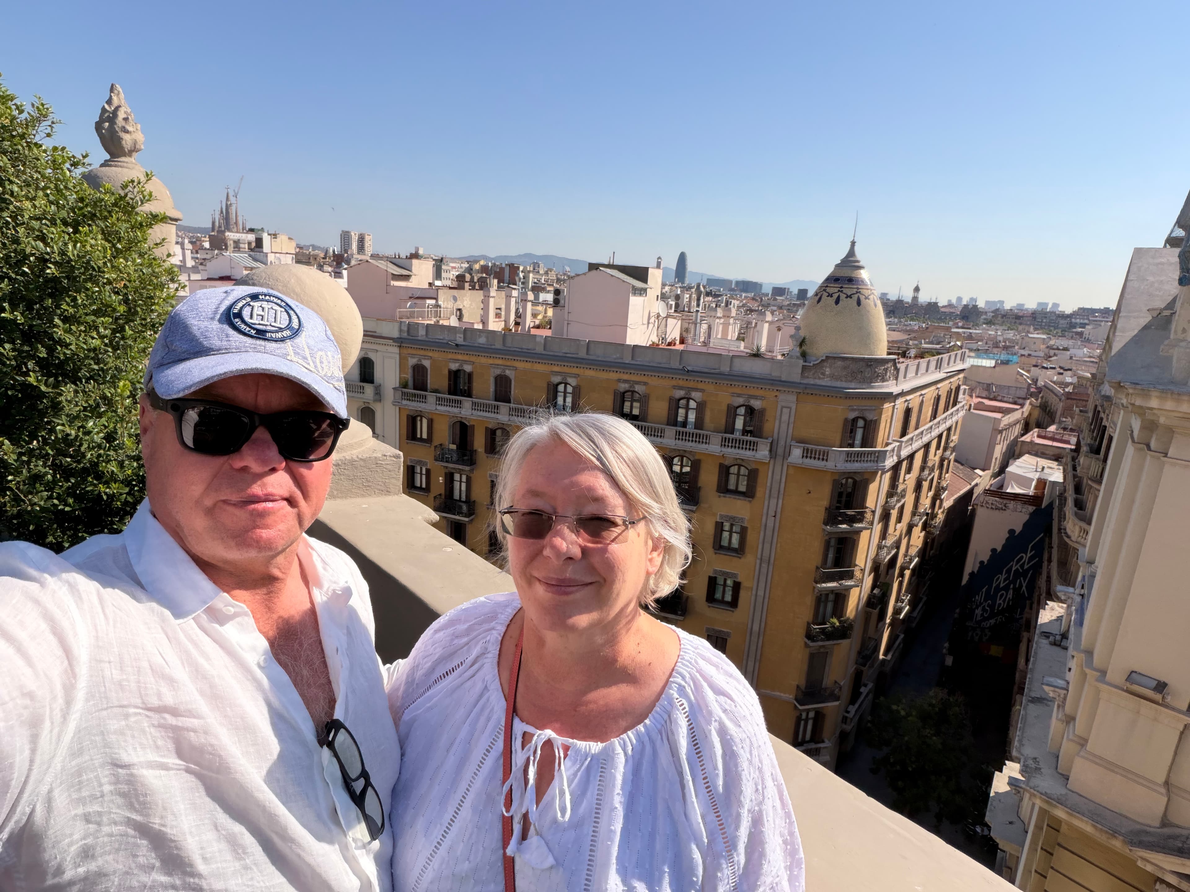 Couple taking a rooftop selfie in Barcelona on a sunny day, with the city skyline, Sagrada Família, and Torre Agbar visible b