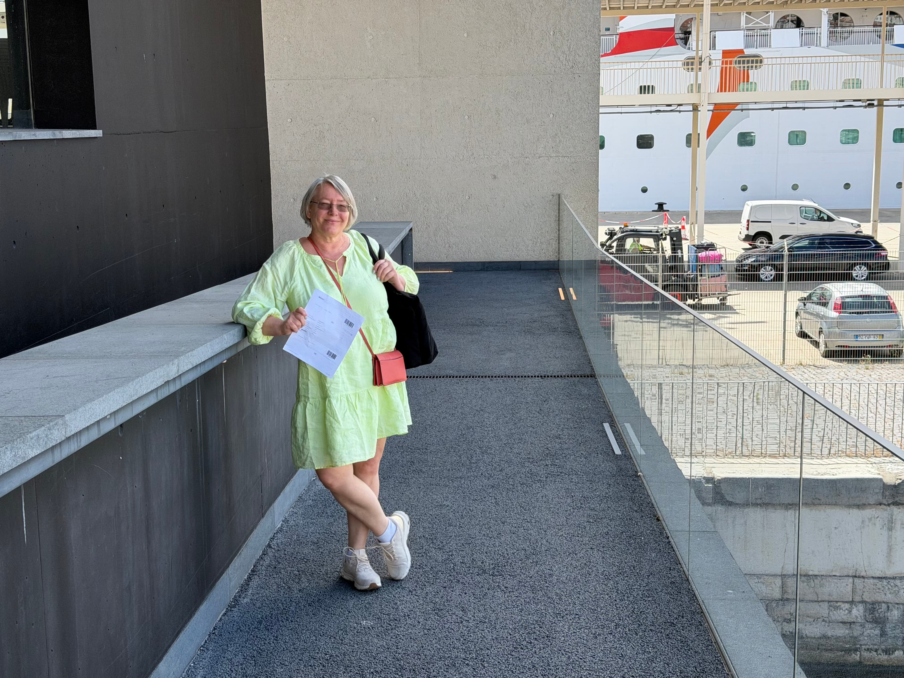 Smiling woman in lime green dress holding boarding documents on a cruise terminal ramp, large white cruise ship visible in ba