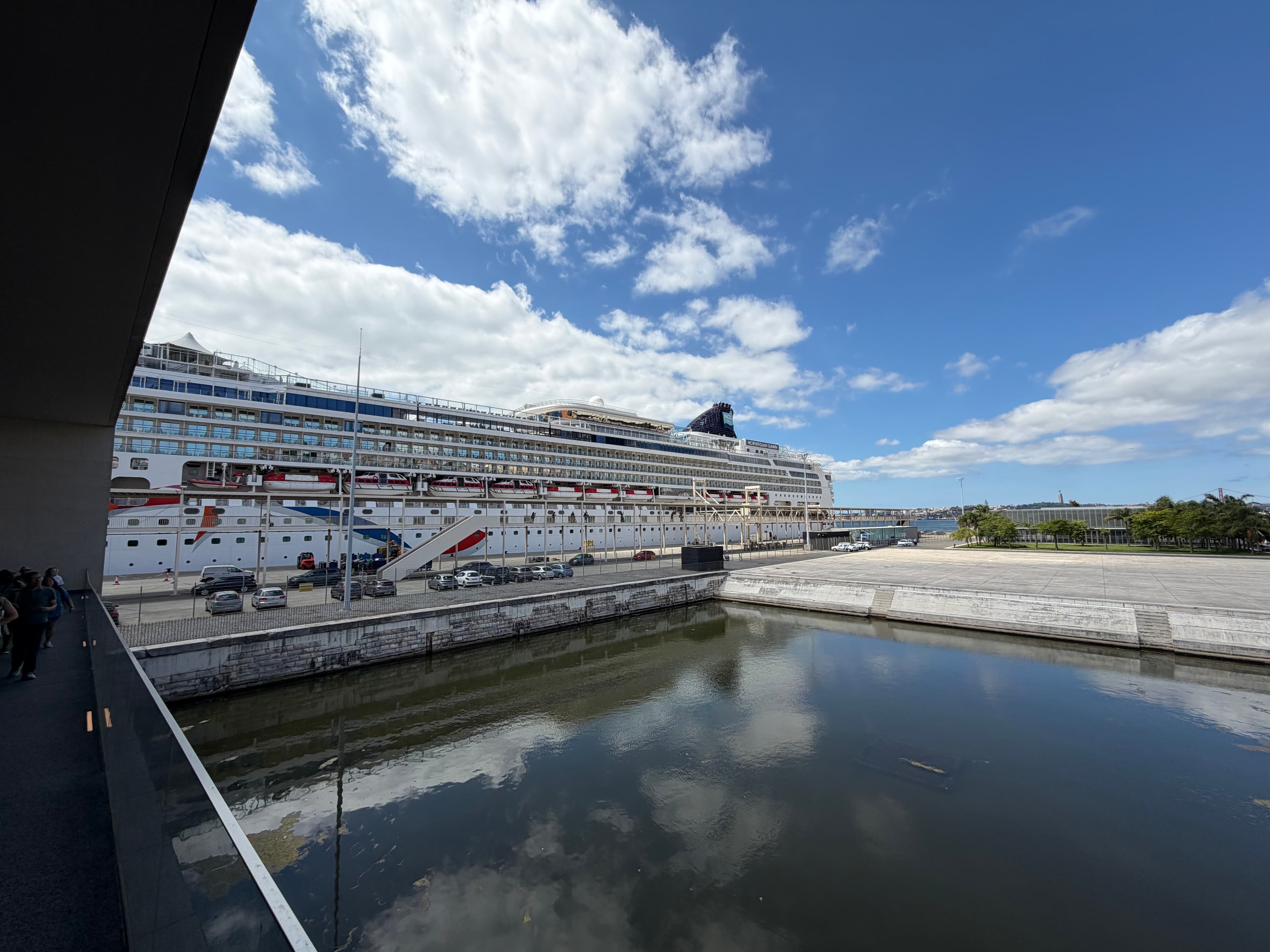 Large white cruise ship docked at a modern port terminal on a sunny day, reflected in calm harbor water below a wide blue sky