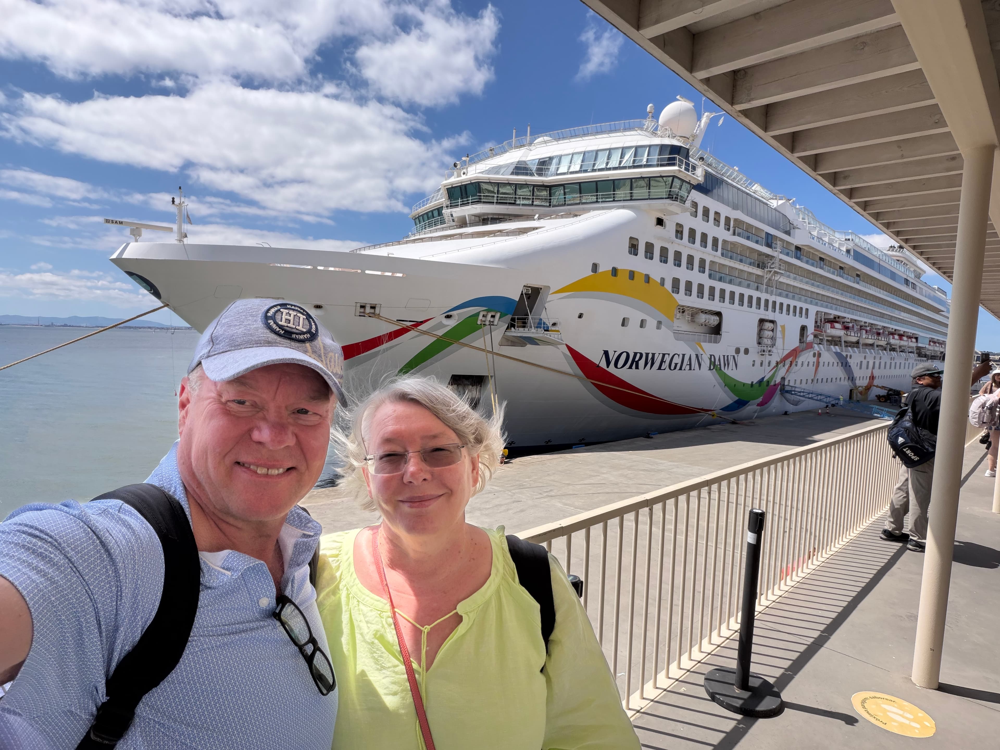 Smiling couple taking a selfie on a sunny cruise terminal dock with the massive white Norwegian Dawn ship docked behind them