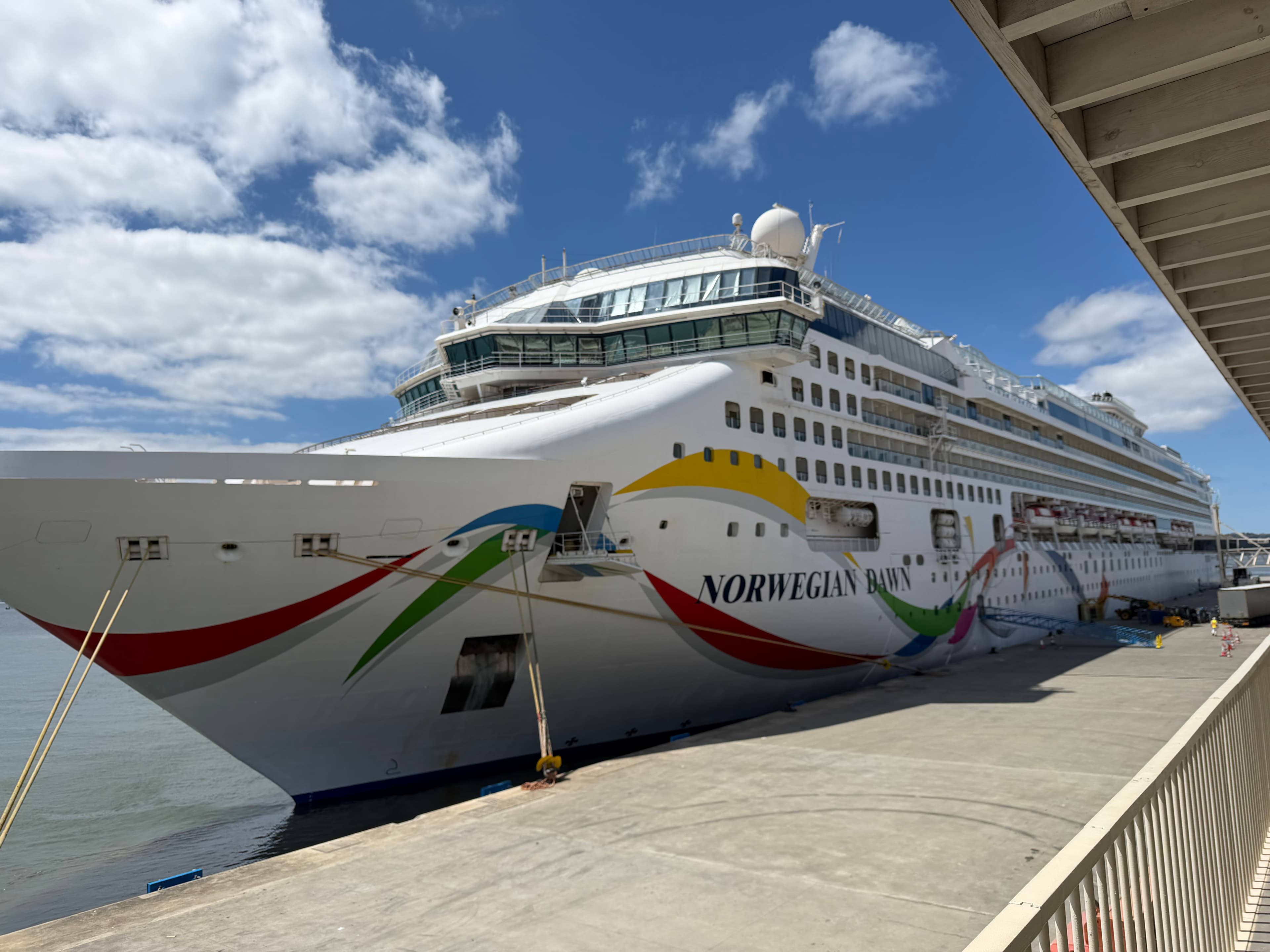 Norwegian Dawn cruise ship docked at port on a sunny day, colorful hull artwork visible against a bright blue sky with scatte
