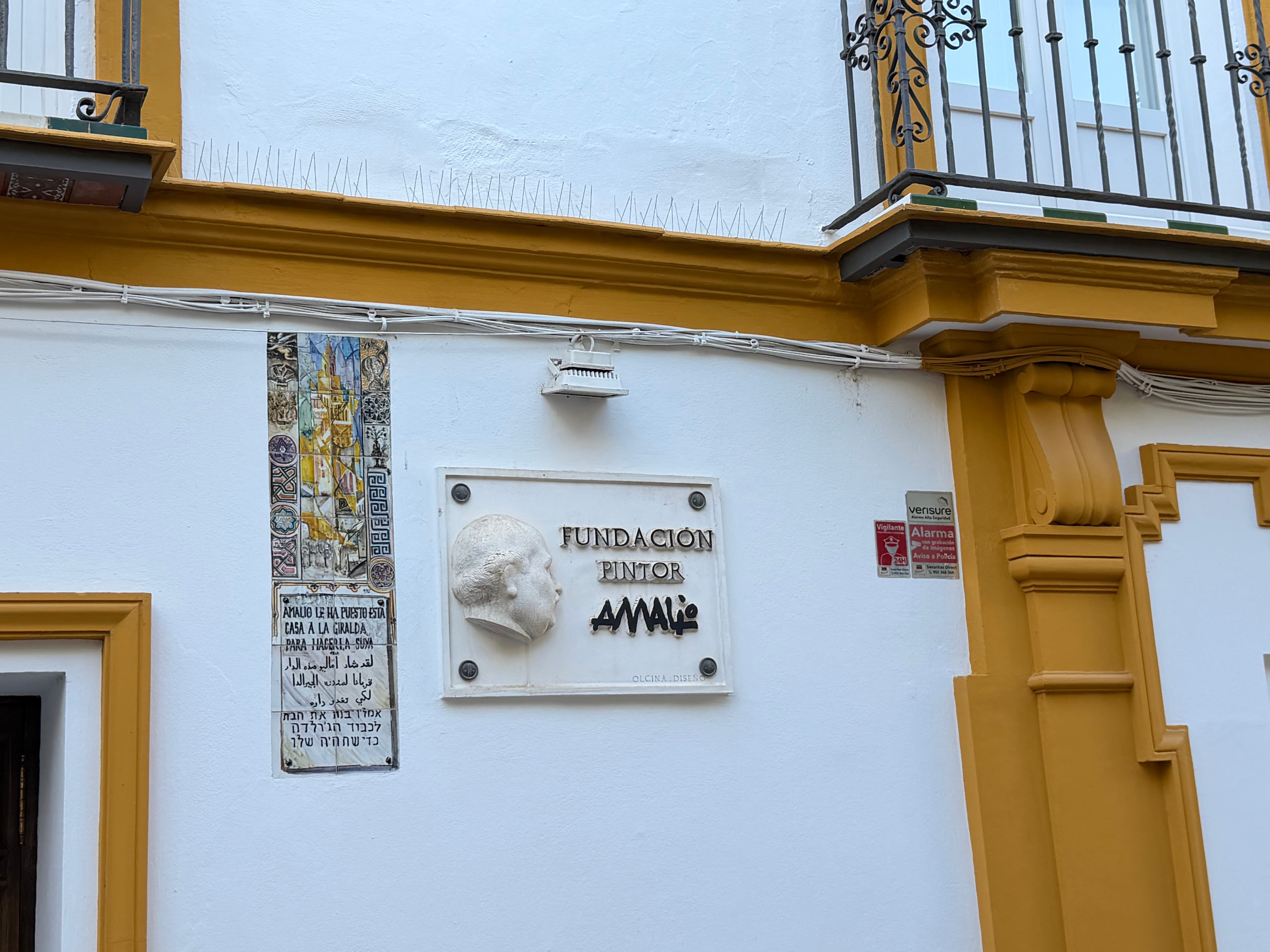 White and ochre Andalusian building facade with a marble plaque for Fundación Pintor Amalio, decorative tile panel, and wroug