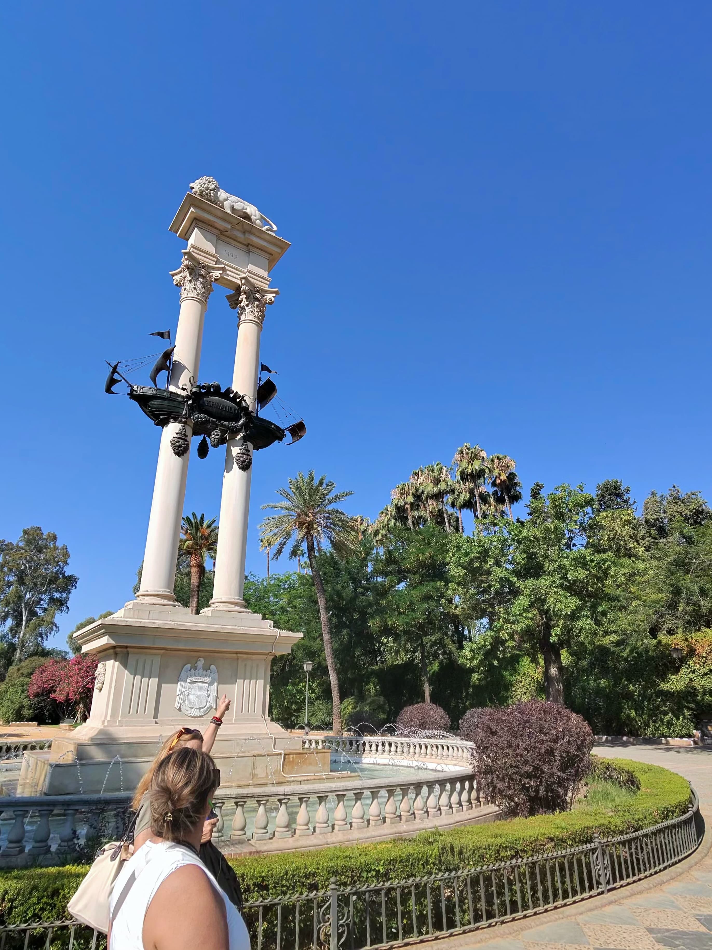 Woman pointing up at the Columbus Monument in Jardines de Murillo, Seville, flanked by palm trees under a vivid blue midday s