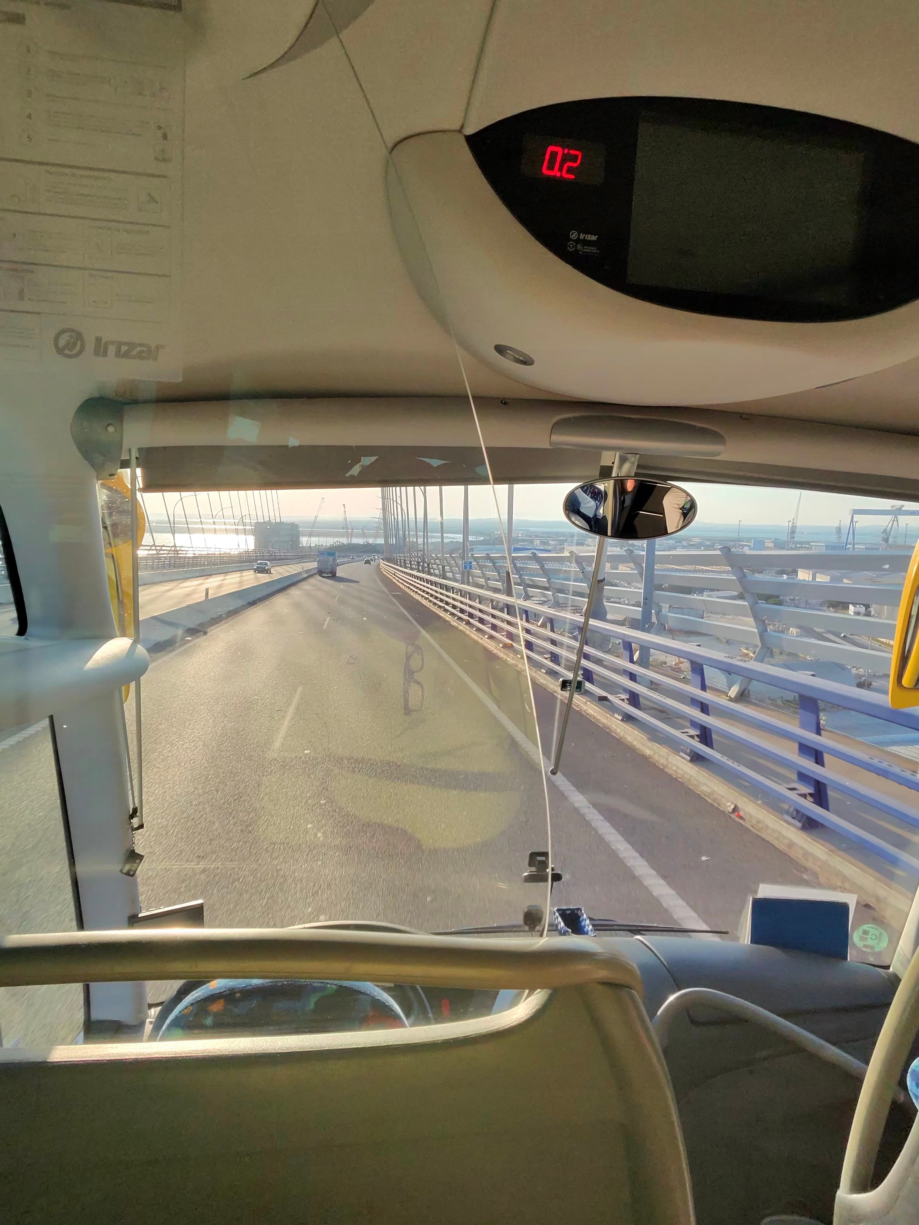 View from front of an Irizar coach crossing a wide cable-stayed bridge over water, daytime with hazy horizon and port cranes