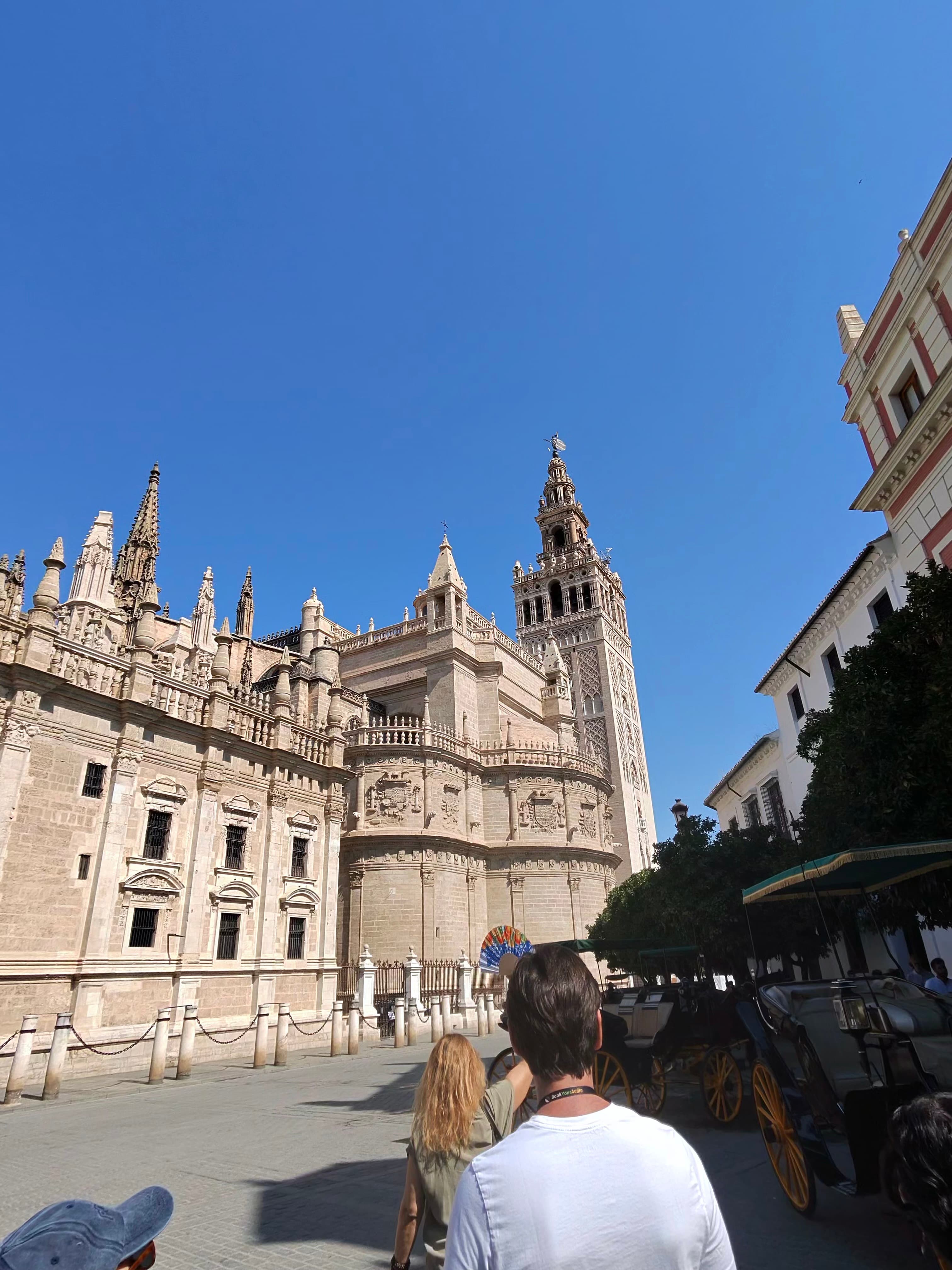 Two tourists gazing up at Seville Cathedral and the Giralda tower under a vivid blue sky, with horse-drawn carriages nearby