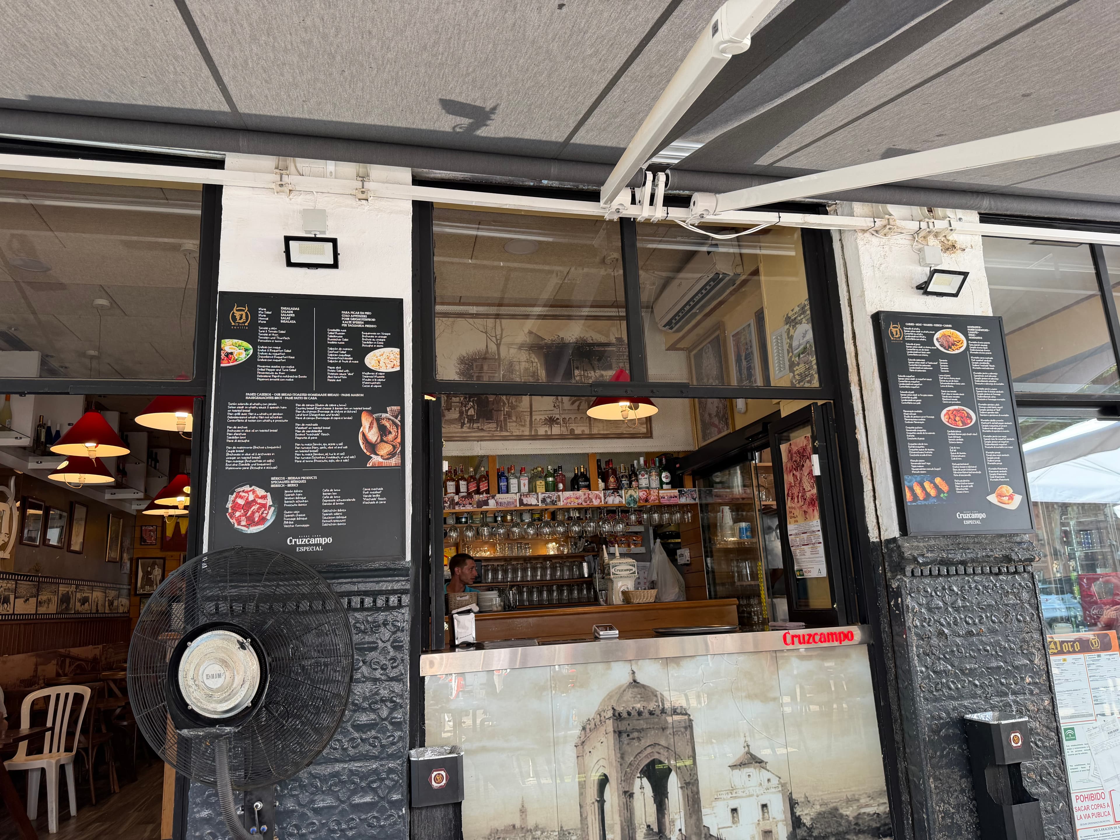 Open-fronted Spanish tapas bar with Cruzcampo branding, black menu boards, a well-stocked bar visible through large windows,