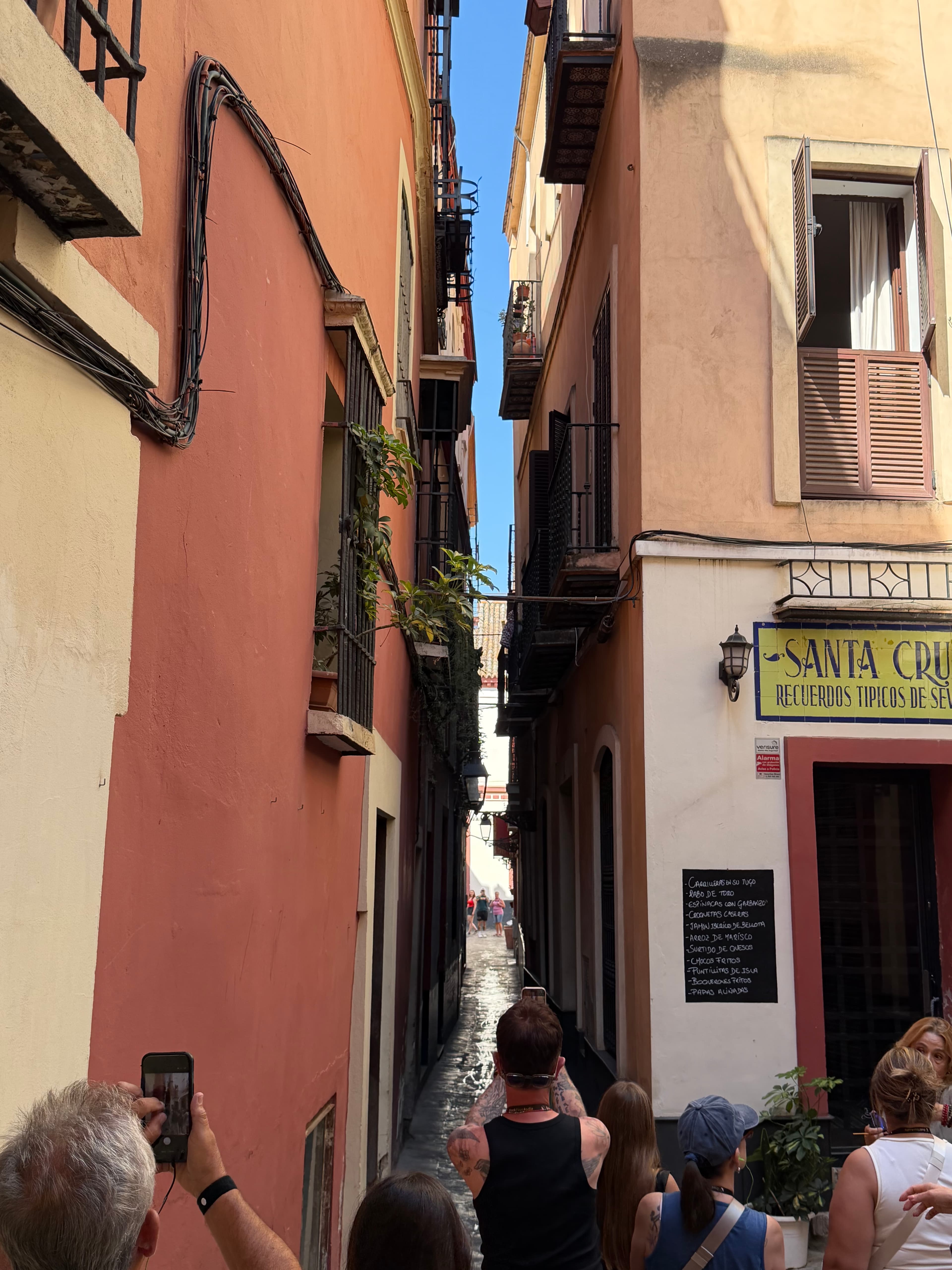 Narrow sunlit alley in Seville's Santa Cruz barrio, terracotta and cream facades with iron balconies, tourists walking under