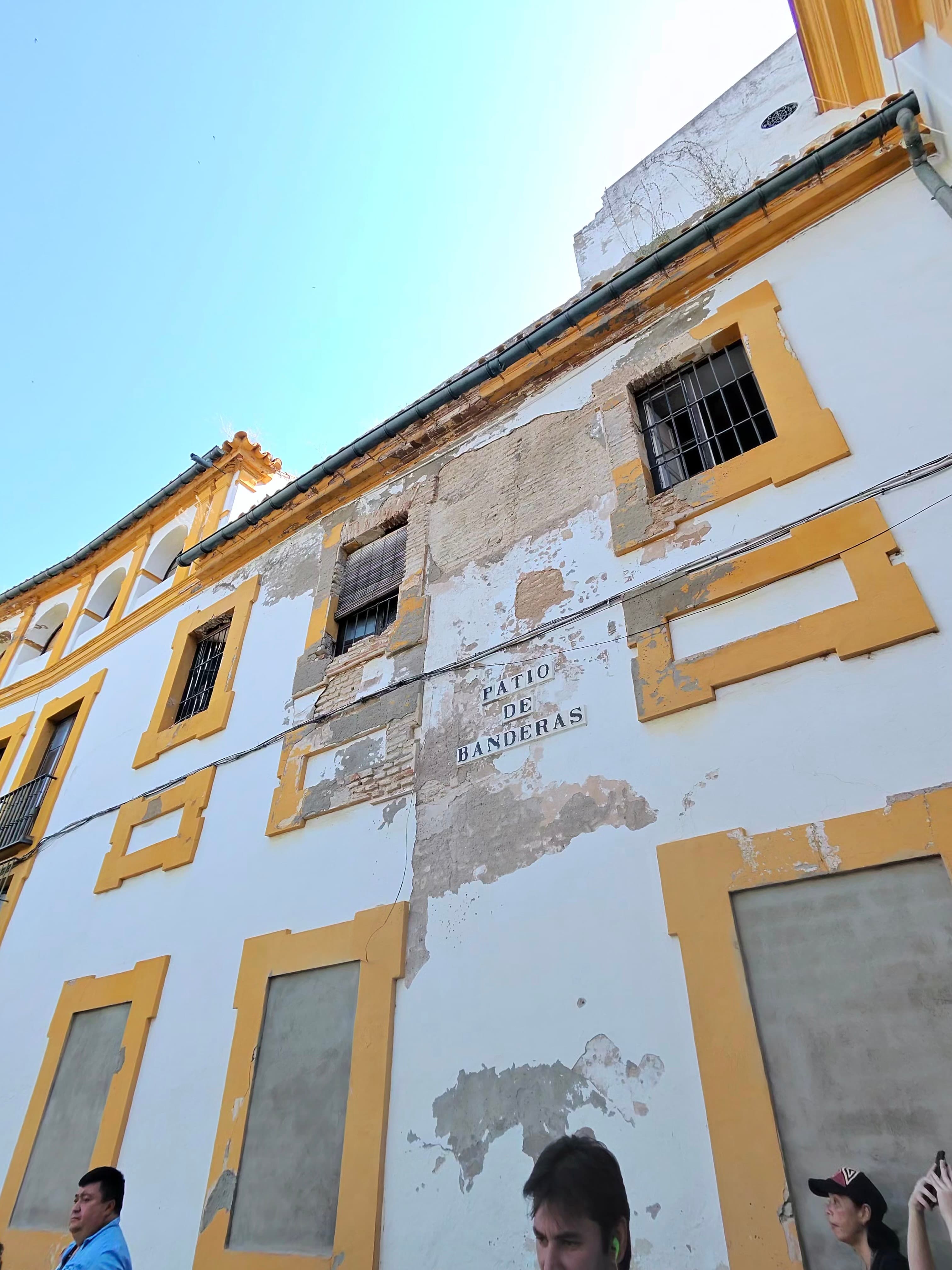 Upward view of weathered white and ochre building facade with 'Patio de Banderas' sign, peeling plaster revealing brick, Sevi