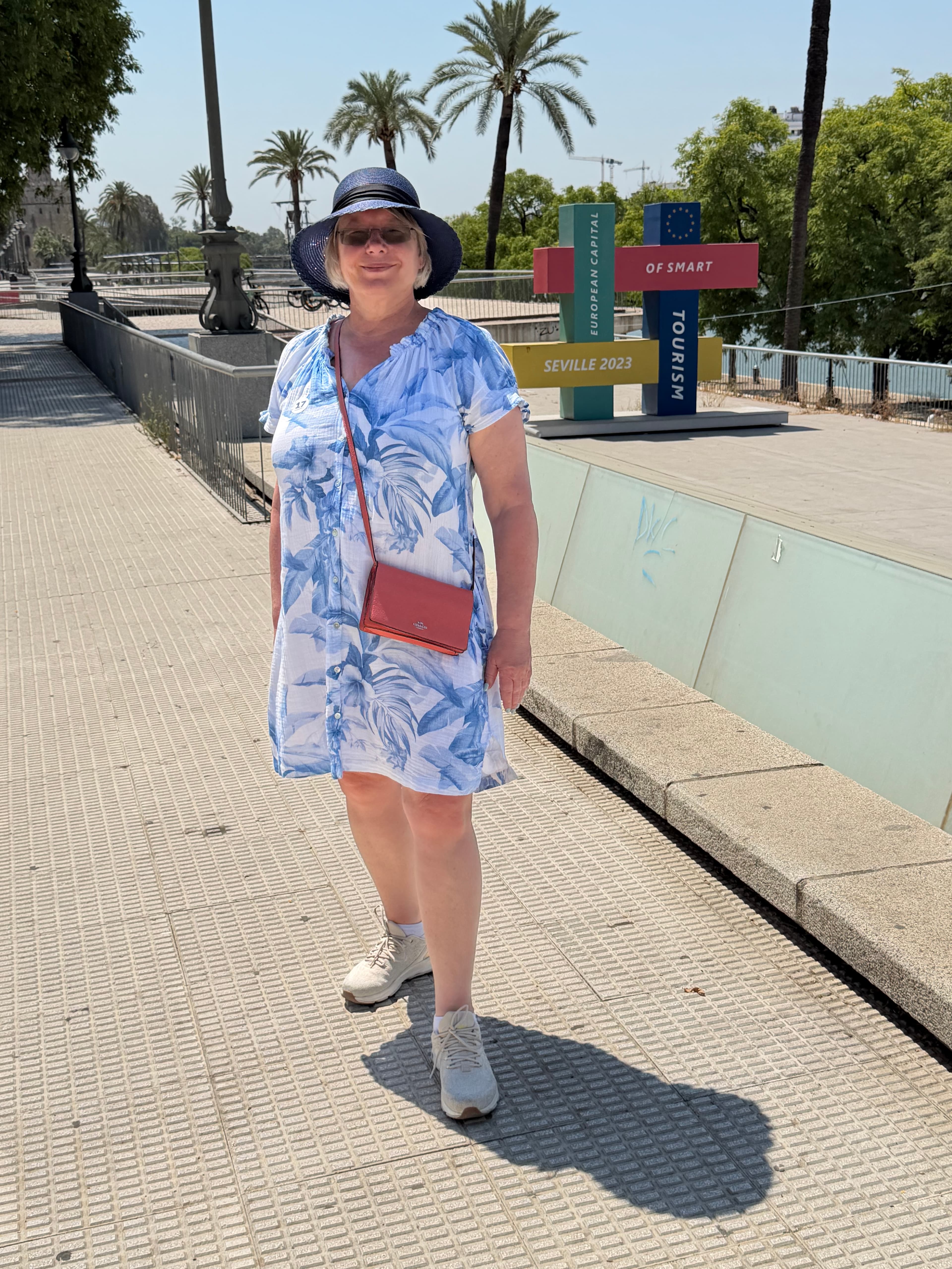 Woman in blue floral dress and sun hat standing on a sunny Seville promenade beside the colorful 'European Capital of Smart T