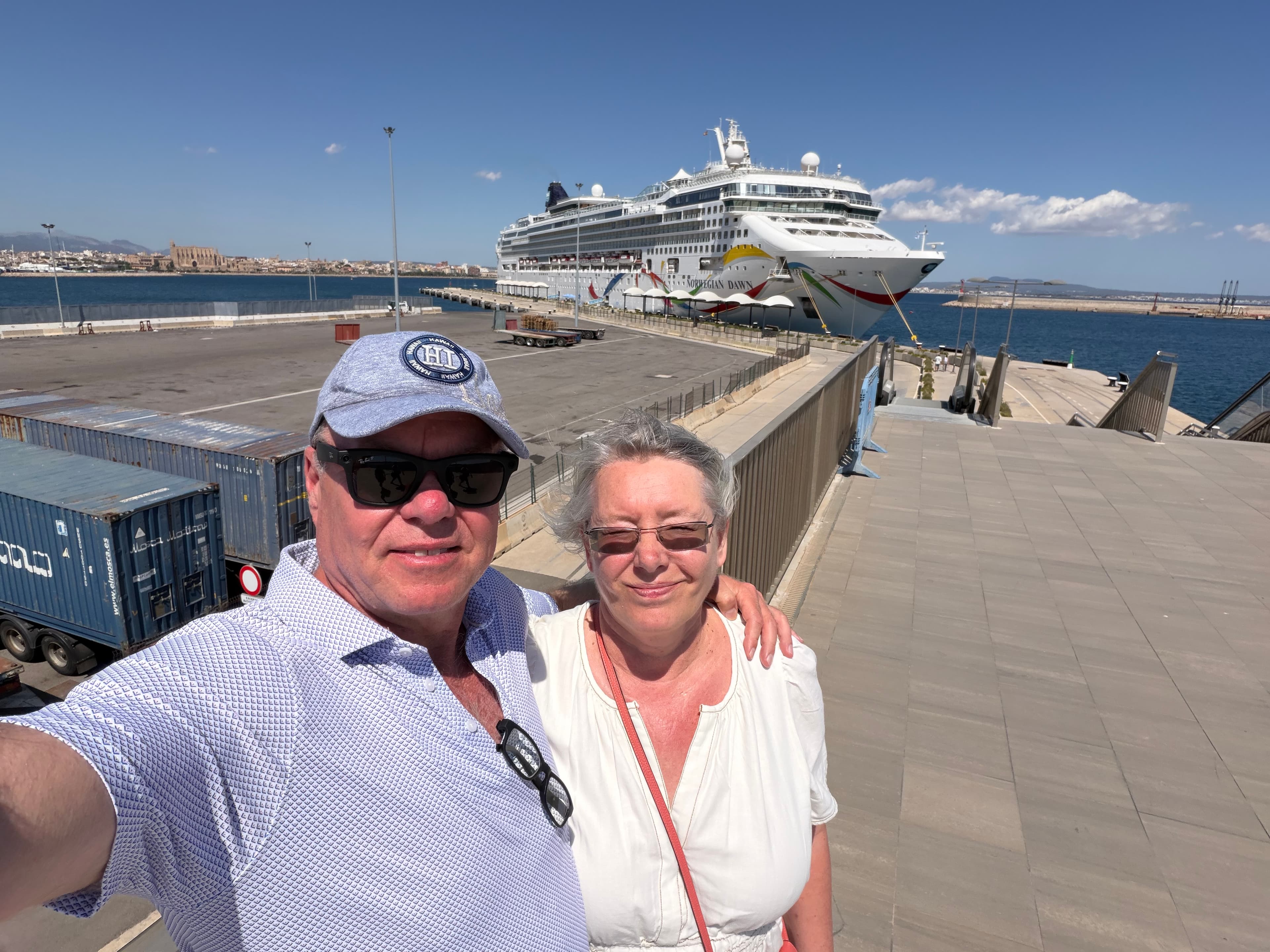 Couple smiling in a sunny port selfie with the Norwegian Dawn cruise ship docked behind them under a clear blue sky