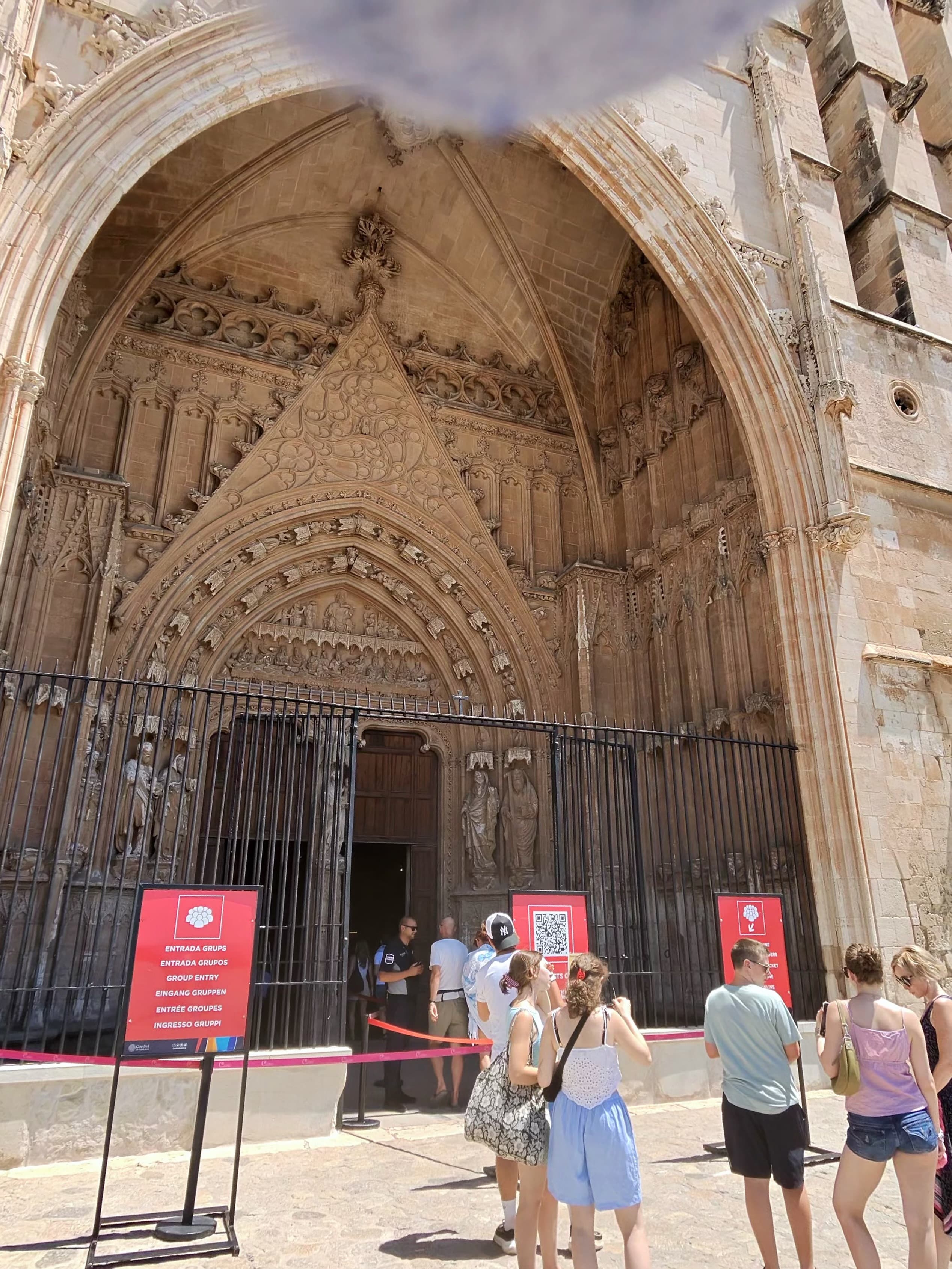 Tourists queuing at the ornate Gothic portal of a Spanish cathedral on a bright sunny day, with intricate stone carvings and