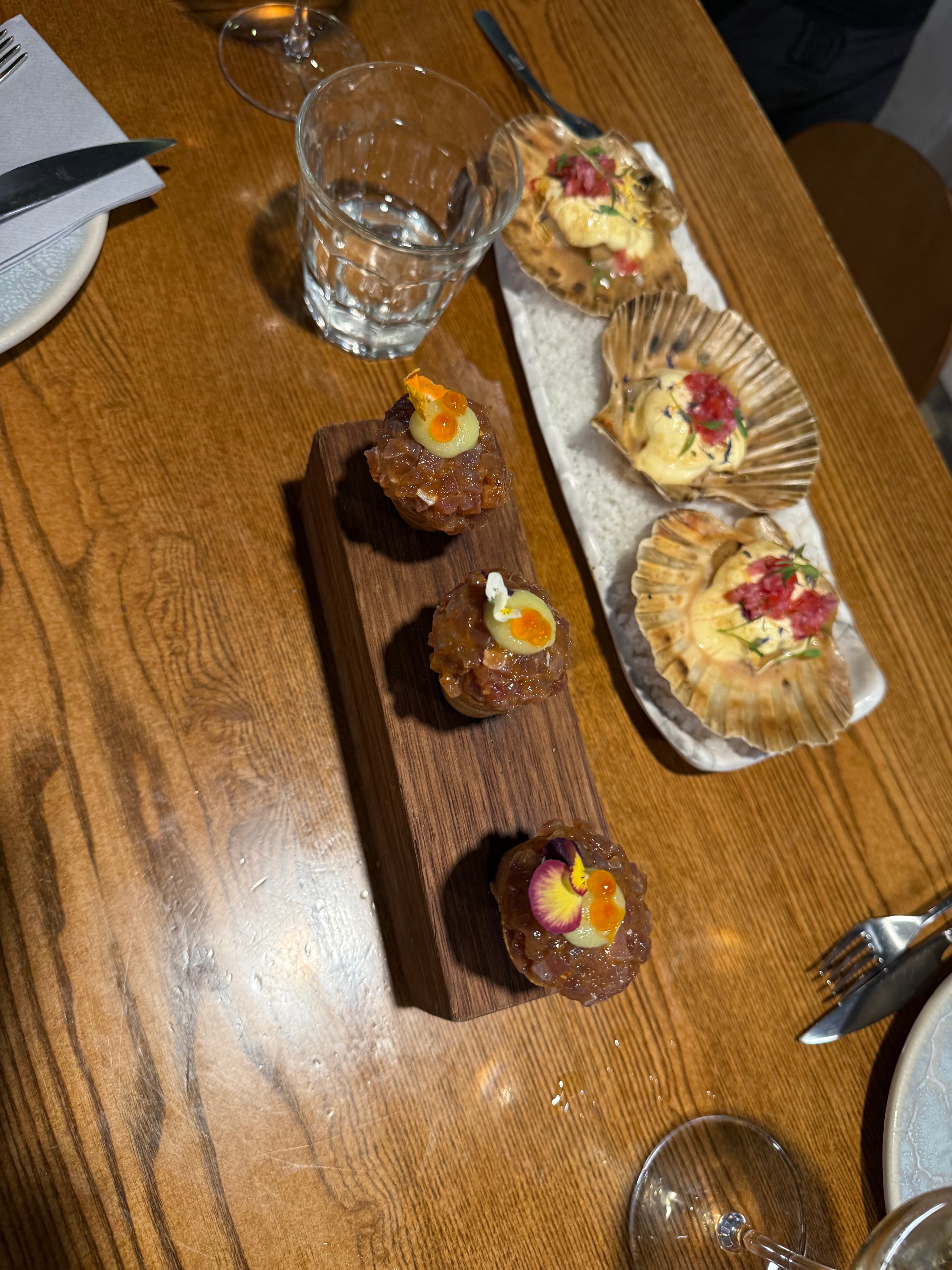 Two tasting courses on a warm oak table: three tartare bites with edible flowers on a wooden board beside four scallops in sh