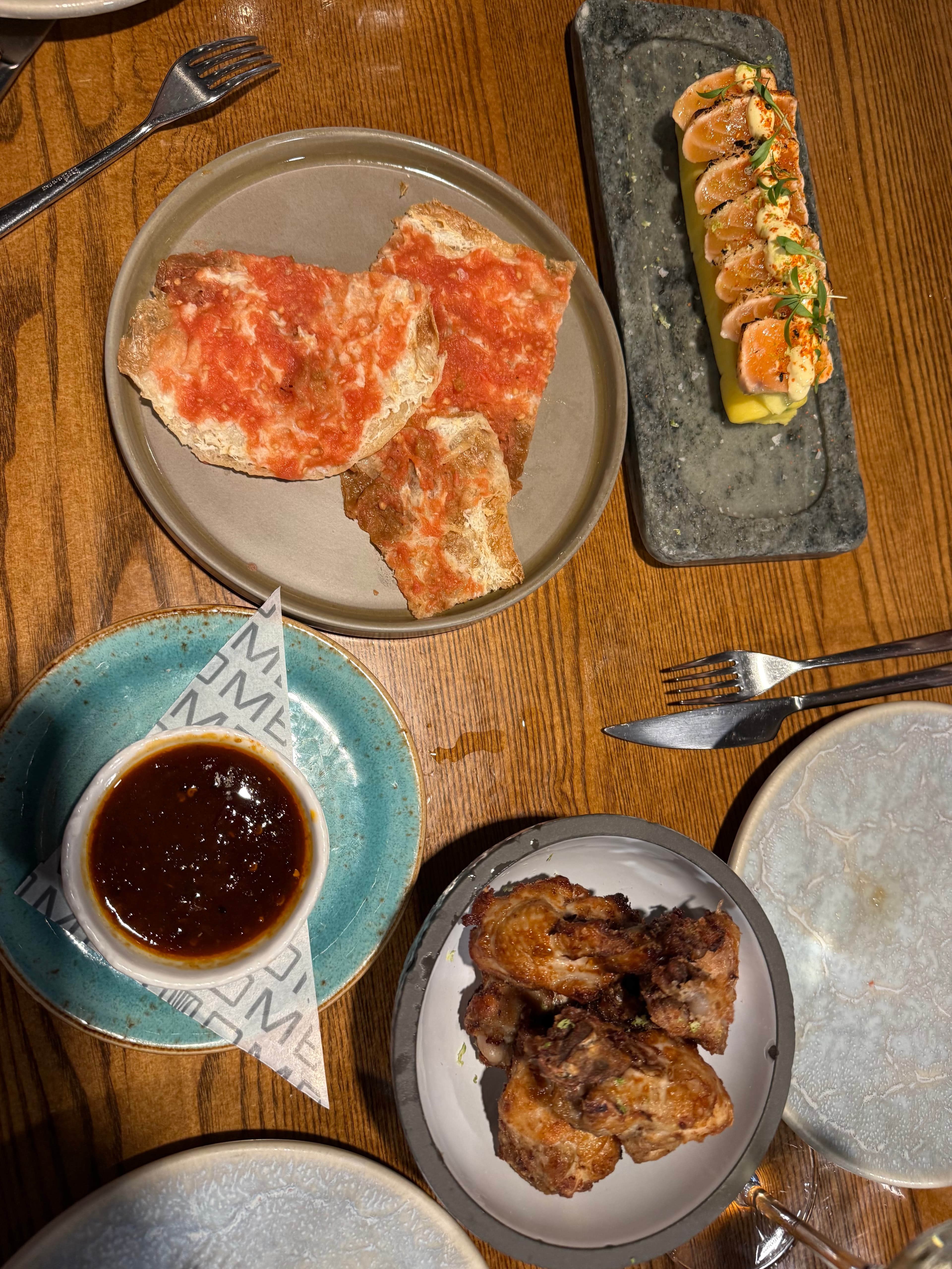 Overhead spread of Spanish tapas on a wooden table: pan con tomate, sliced salmon on a slate board, fried meat bites, and a d
