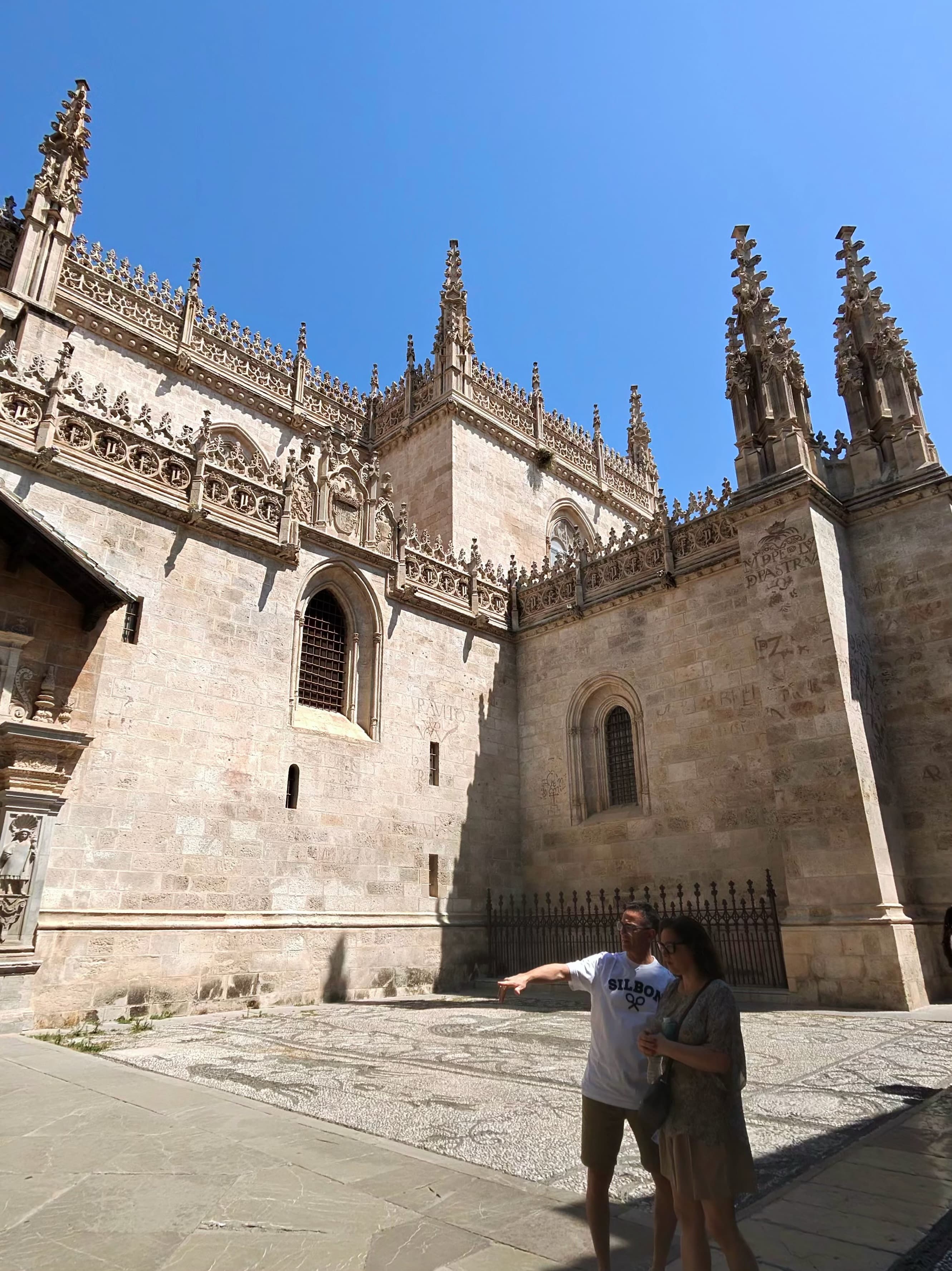 Two tourists pointing and exploring the ornate Isabelline Gothic exterior of Granada's Royal Chapel under a vivid blue midday