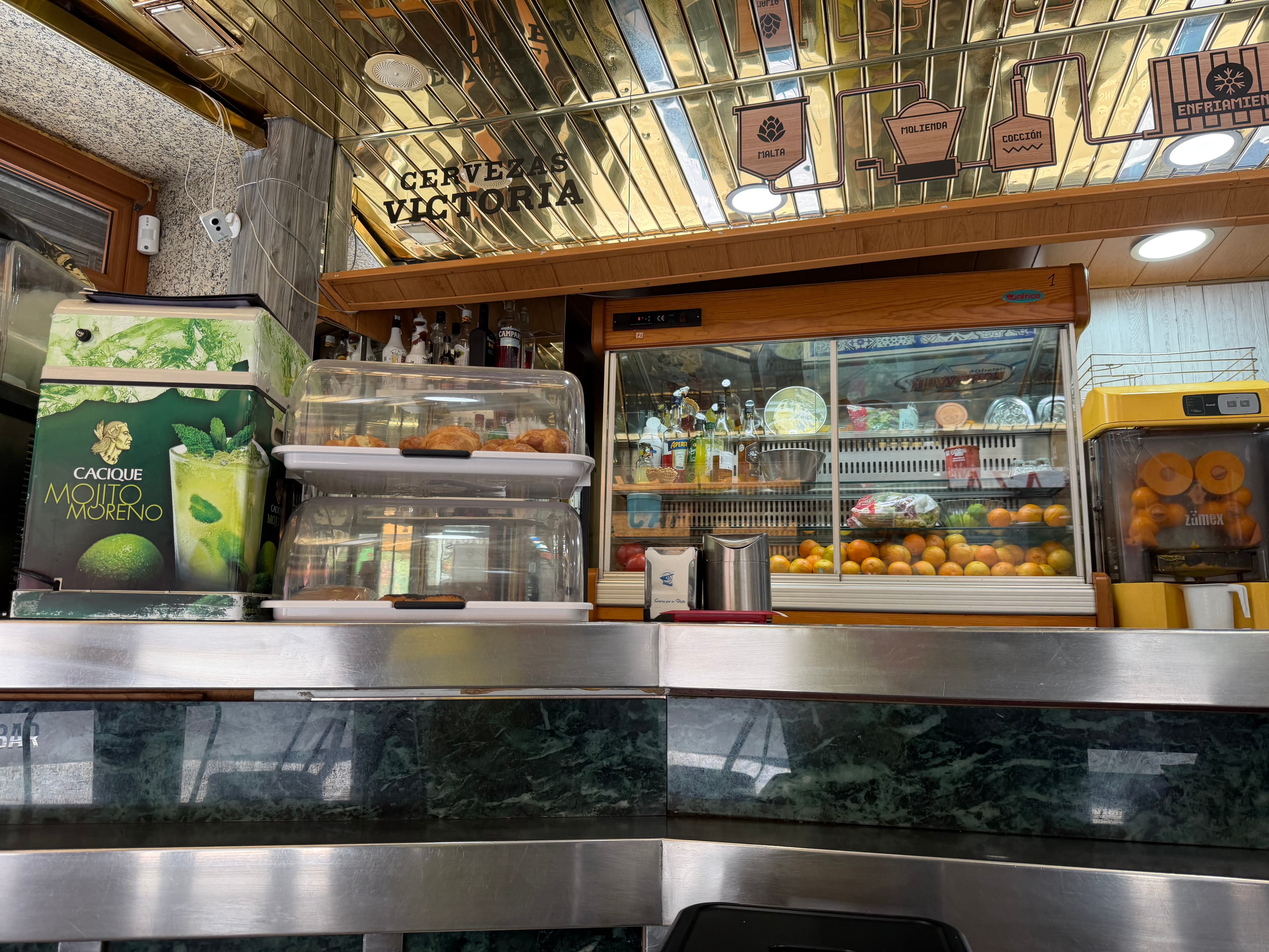 Spanish bar counter with Cervezas Victoria signage, pastry display cases, fresh oranges for juicing, and a mirrored gold ceil