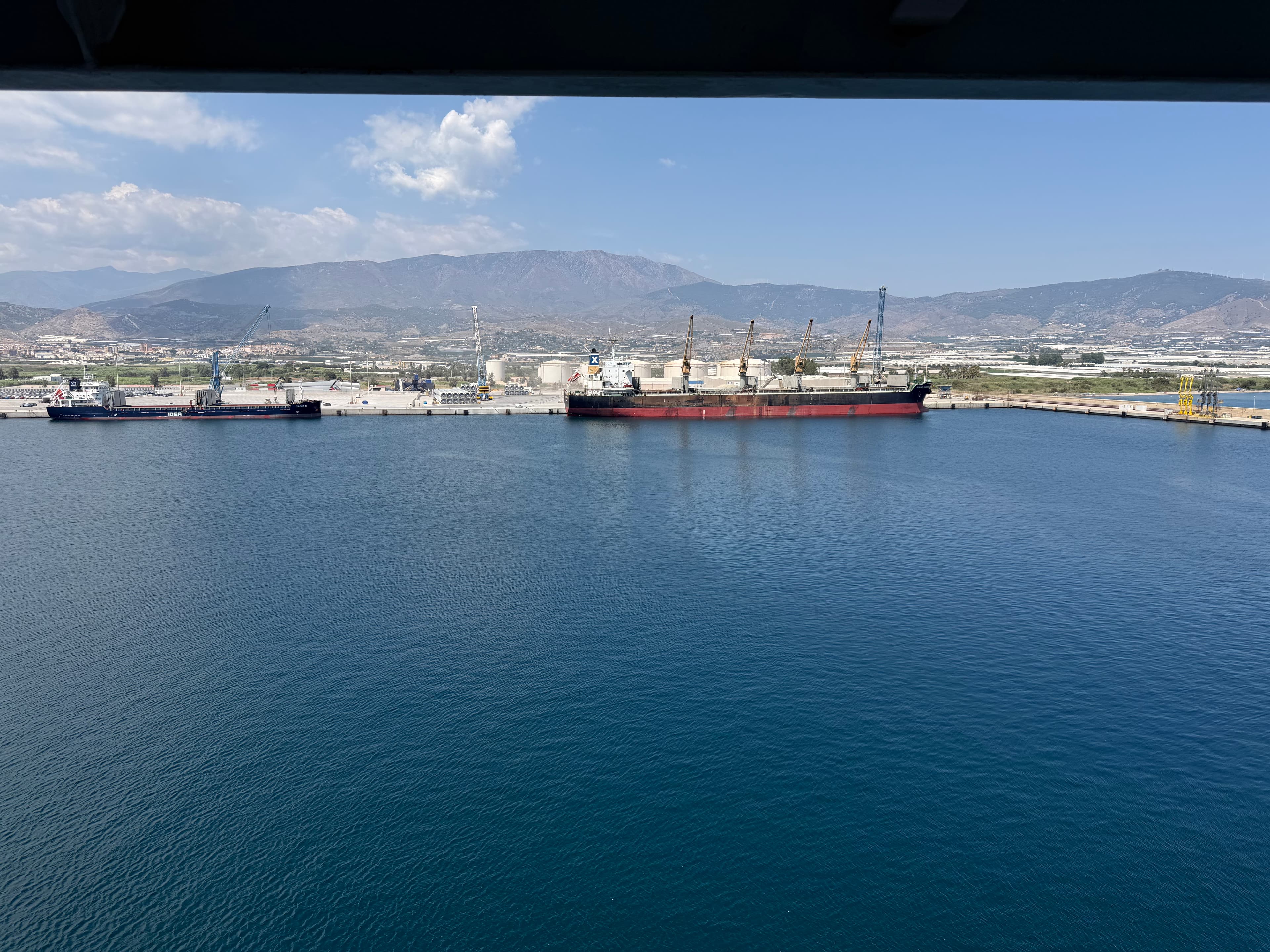 Two large cargo ships docked at a Mediterranean port on a sunny day, with industrial cranes and arid mountains stretching acr