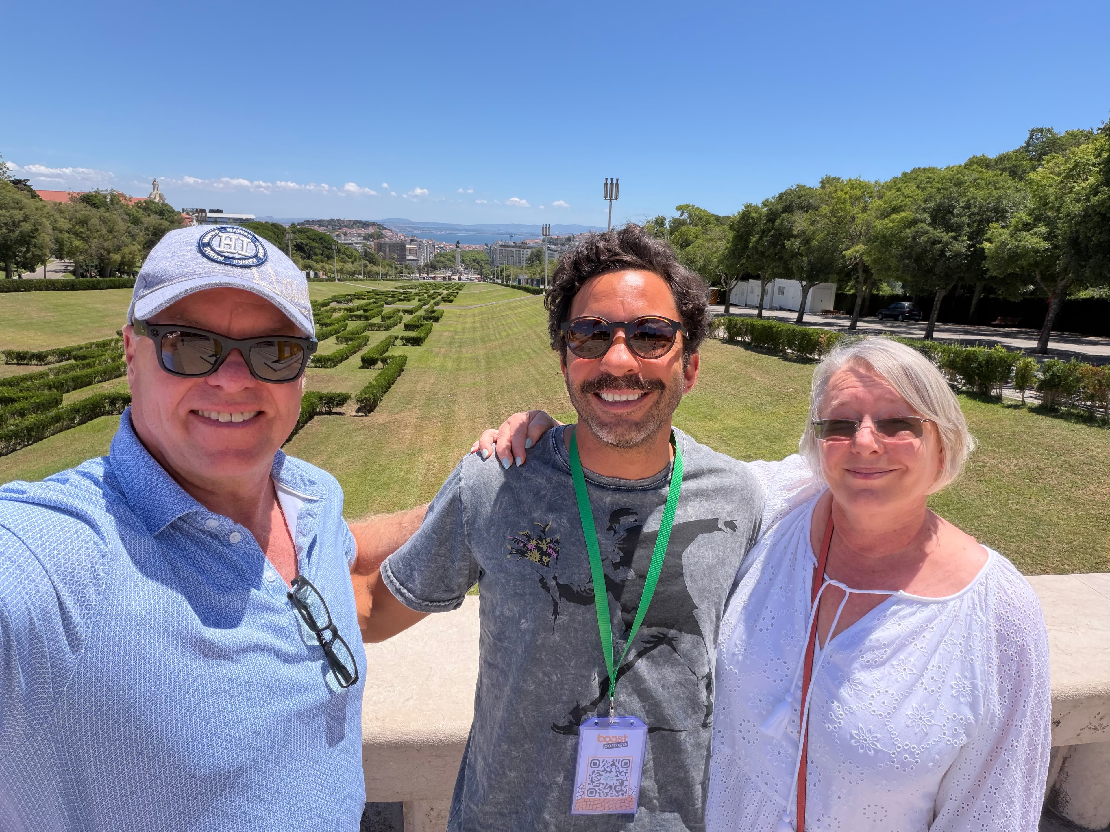 Three smiling travelers posing at a sun-drenched overlook above Parque Eduardo VII in Lisbon, with manicured hedgerows and th