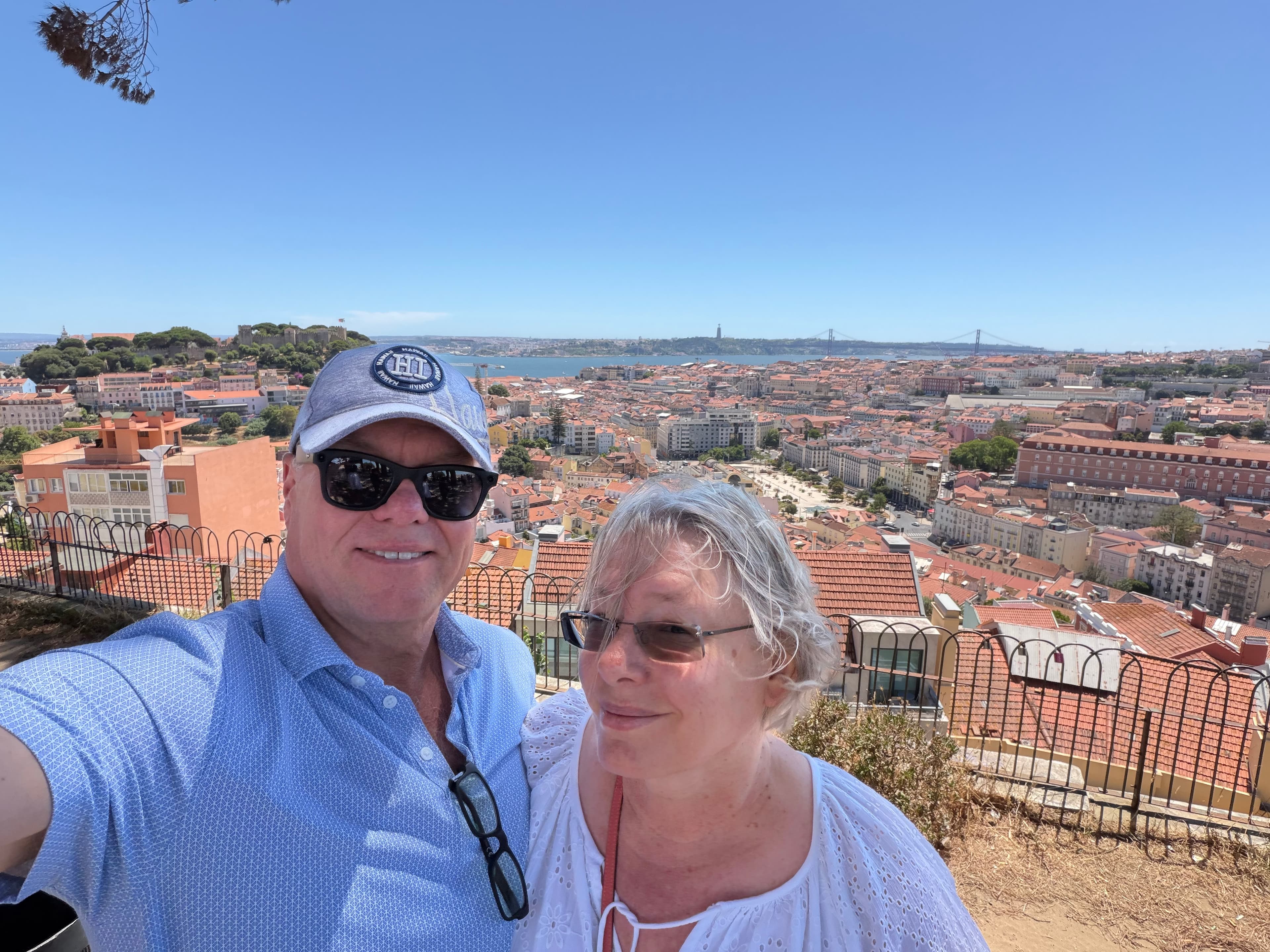 Couple taking a sunny selfie at a Lisbon hilltop viewpoint, with terracotta rooftops, the Tagus River, and the 25 de Abril Br
