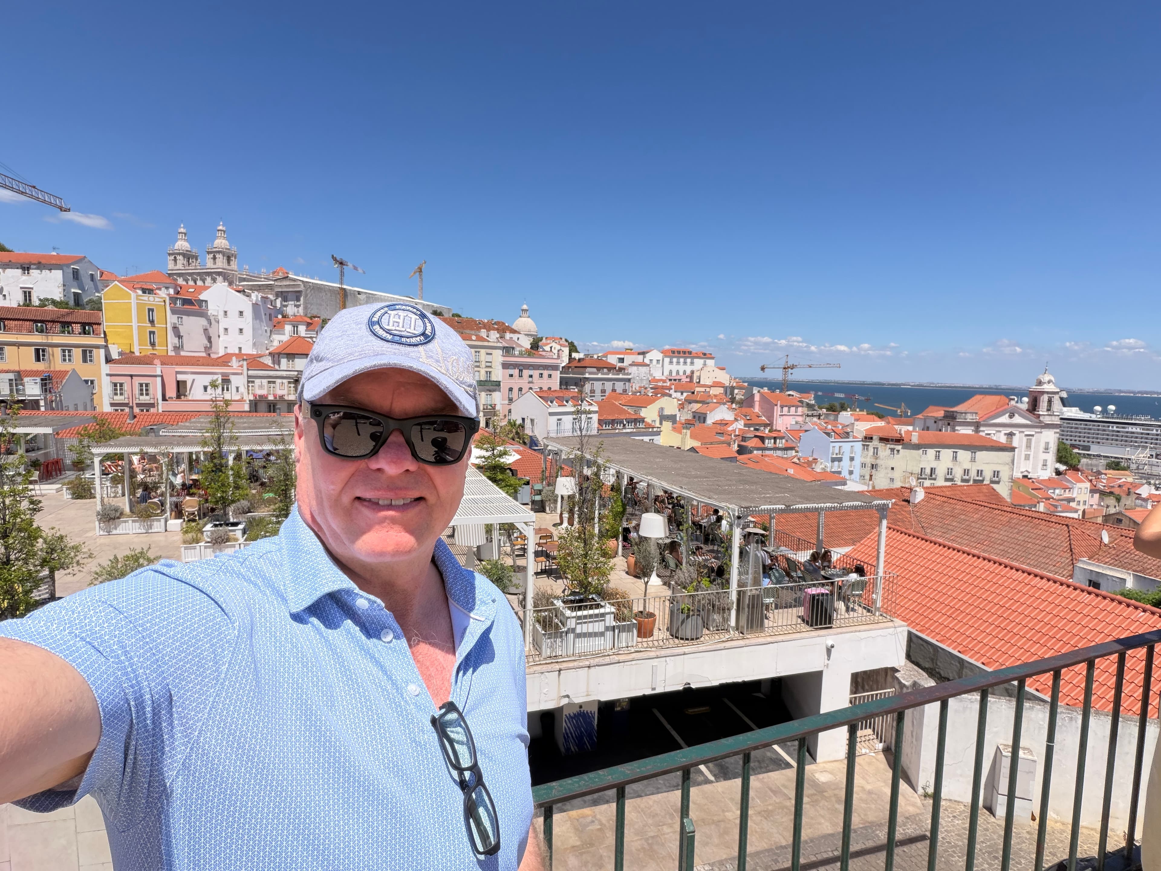 Smiling traveler taking a selfie at a Lisbon miradouro overlook, red-tiled rooftops and the Tagus River stretching behind him