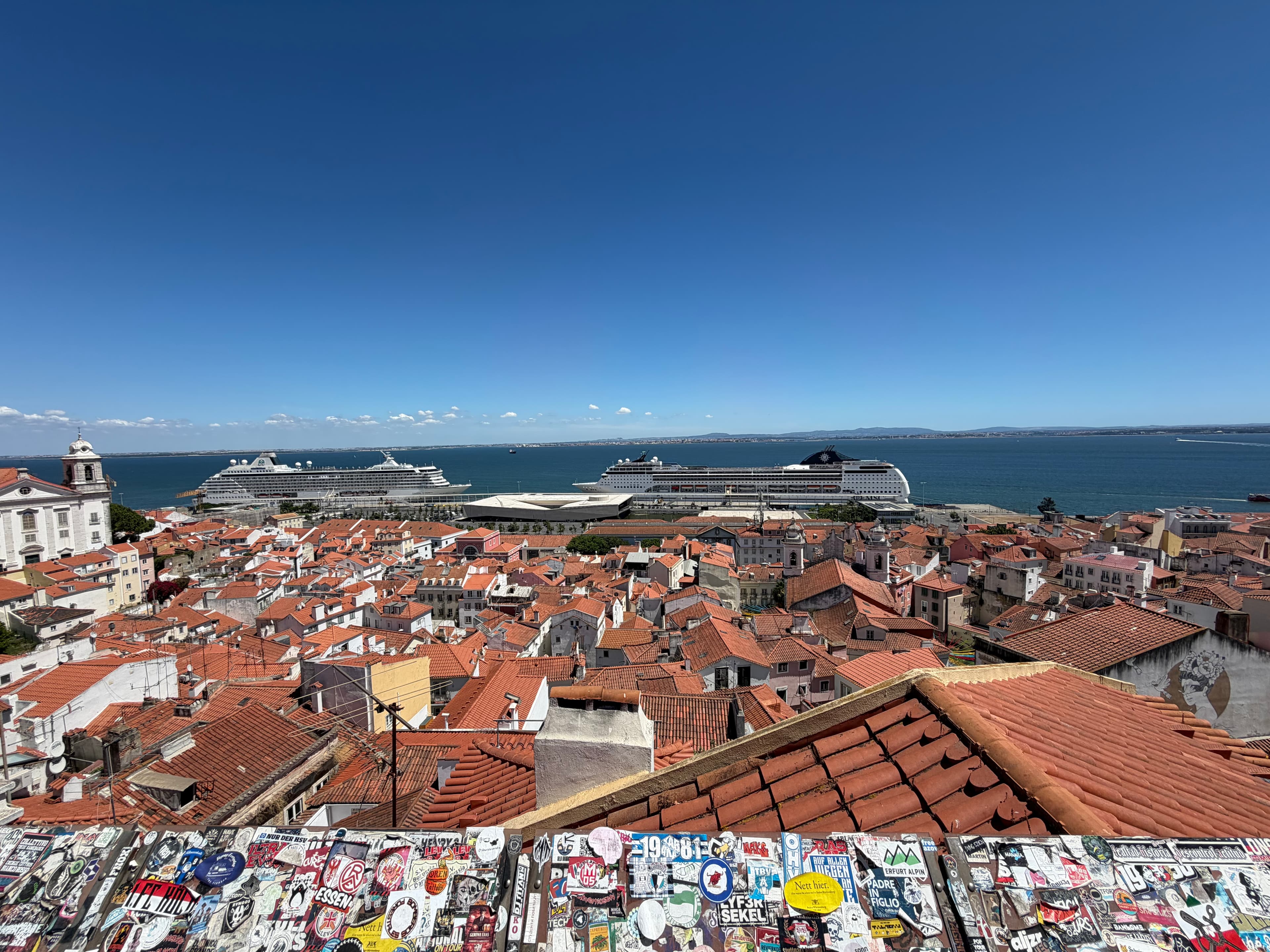 Lisbon's Alfama rooftops of terracotta tiles stretch toward the Tagus River where two cruise ships are docked, under a vivid