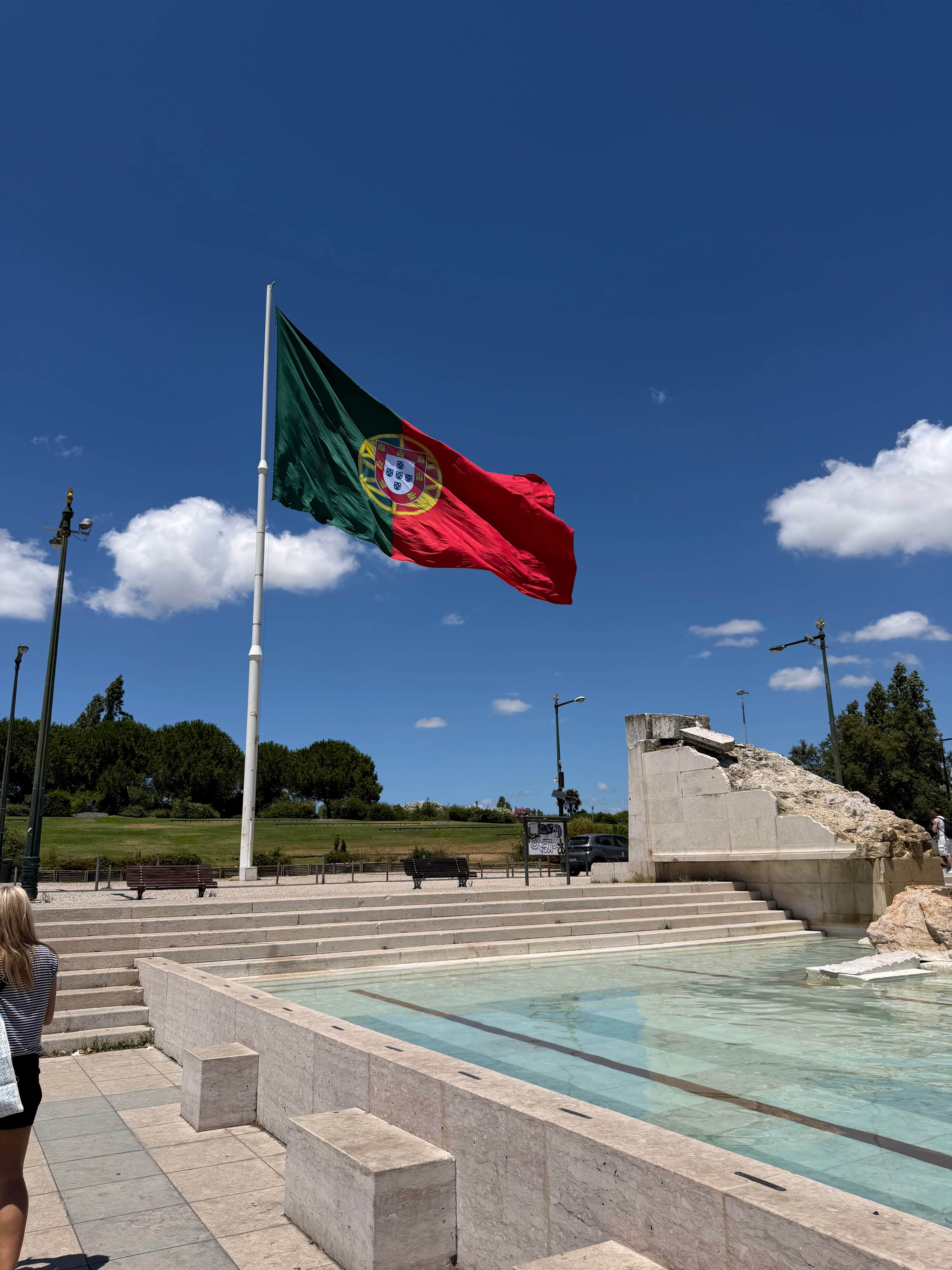 Large Portuguese flag billowing in the wind above a marble fountain plaza under a vivid blue midday sky in Lisbon