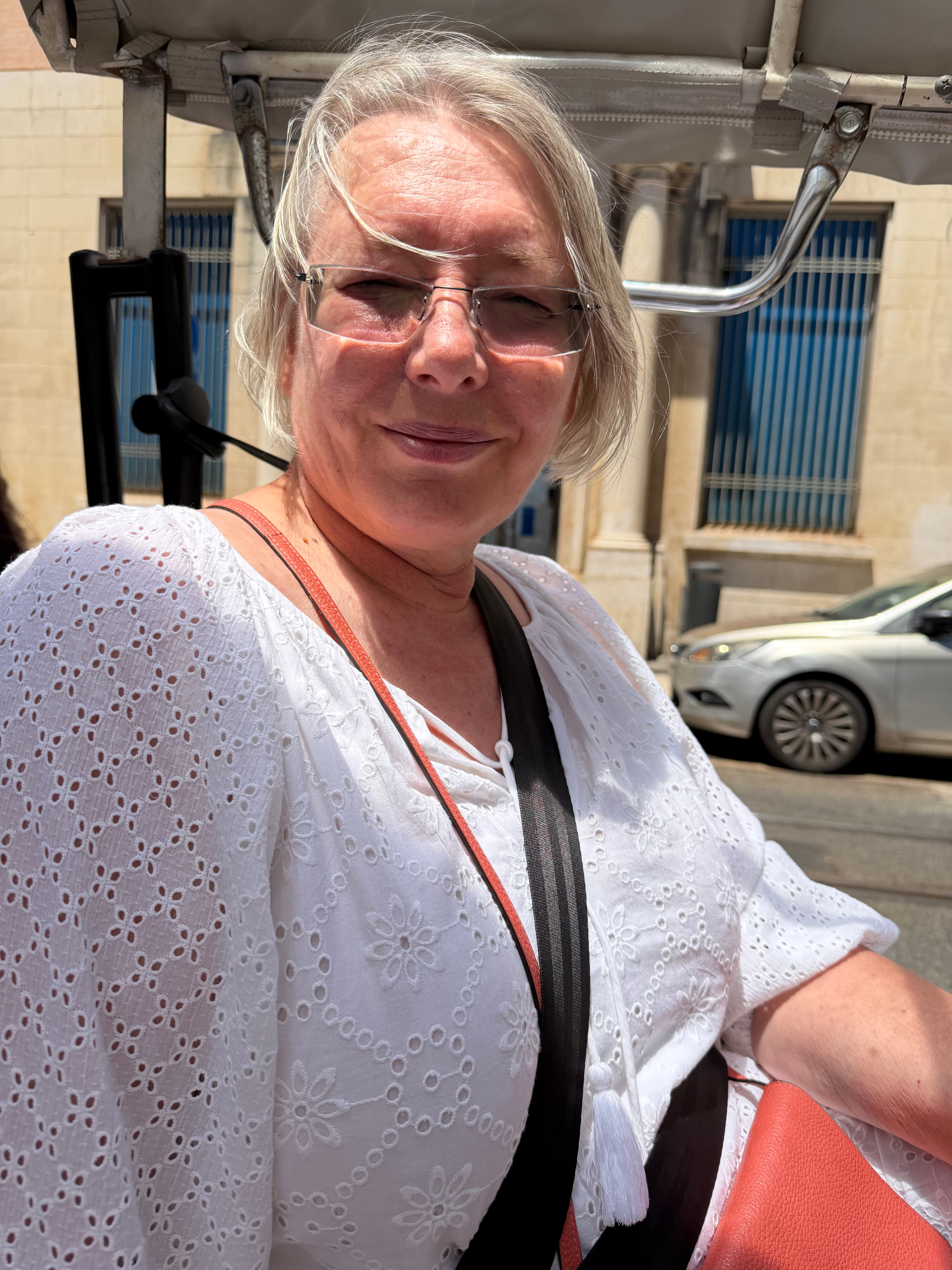 Silver-haired woman in white eyelet blouse and glasses smiling in bright midday sun, sandy limestone buildings visible behind