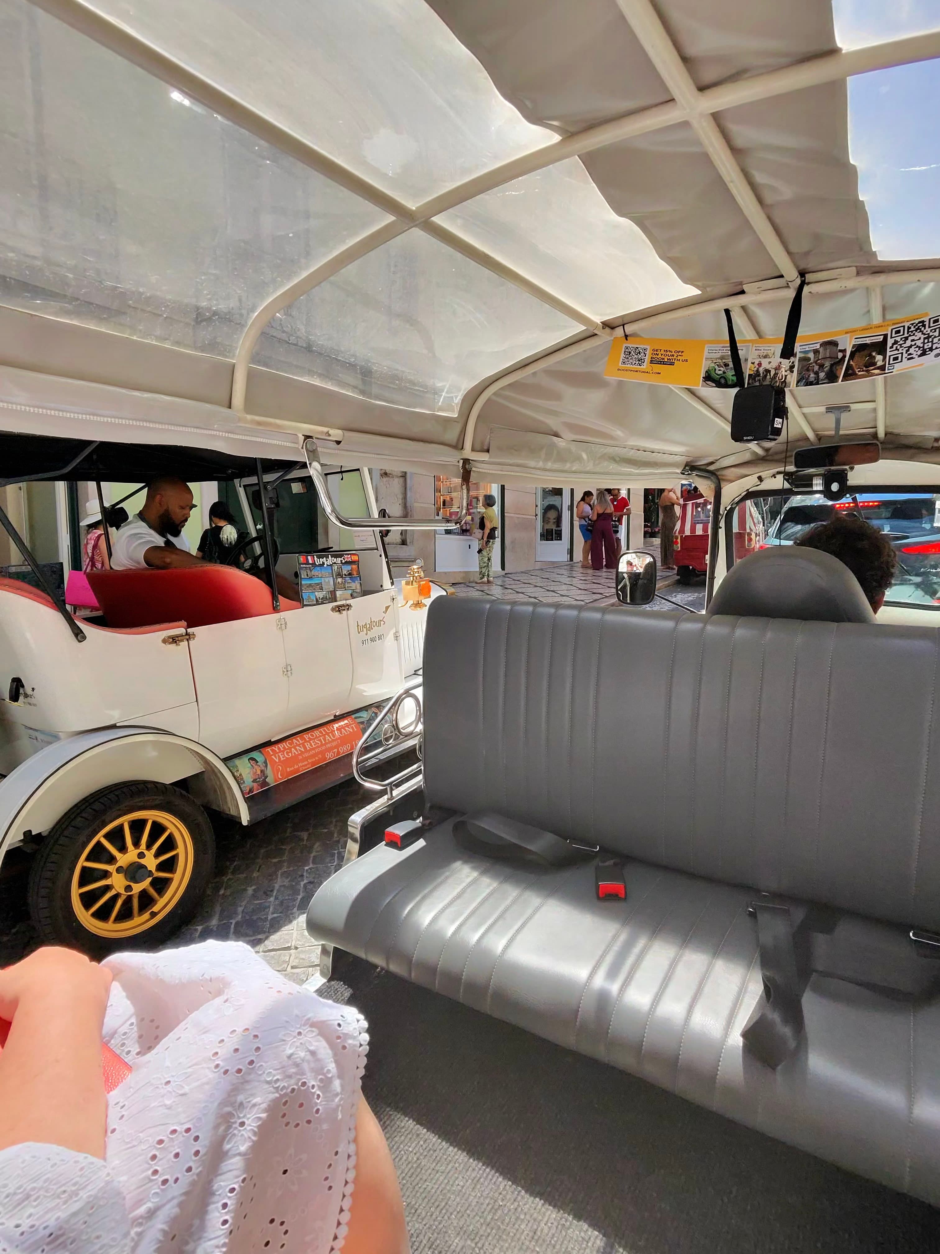 Interior of an open-air tuk-tuk tour vehicle stopped on a cobblestone street, with a white canopy roof, gray bench seats, and