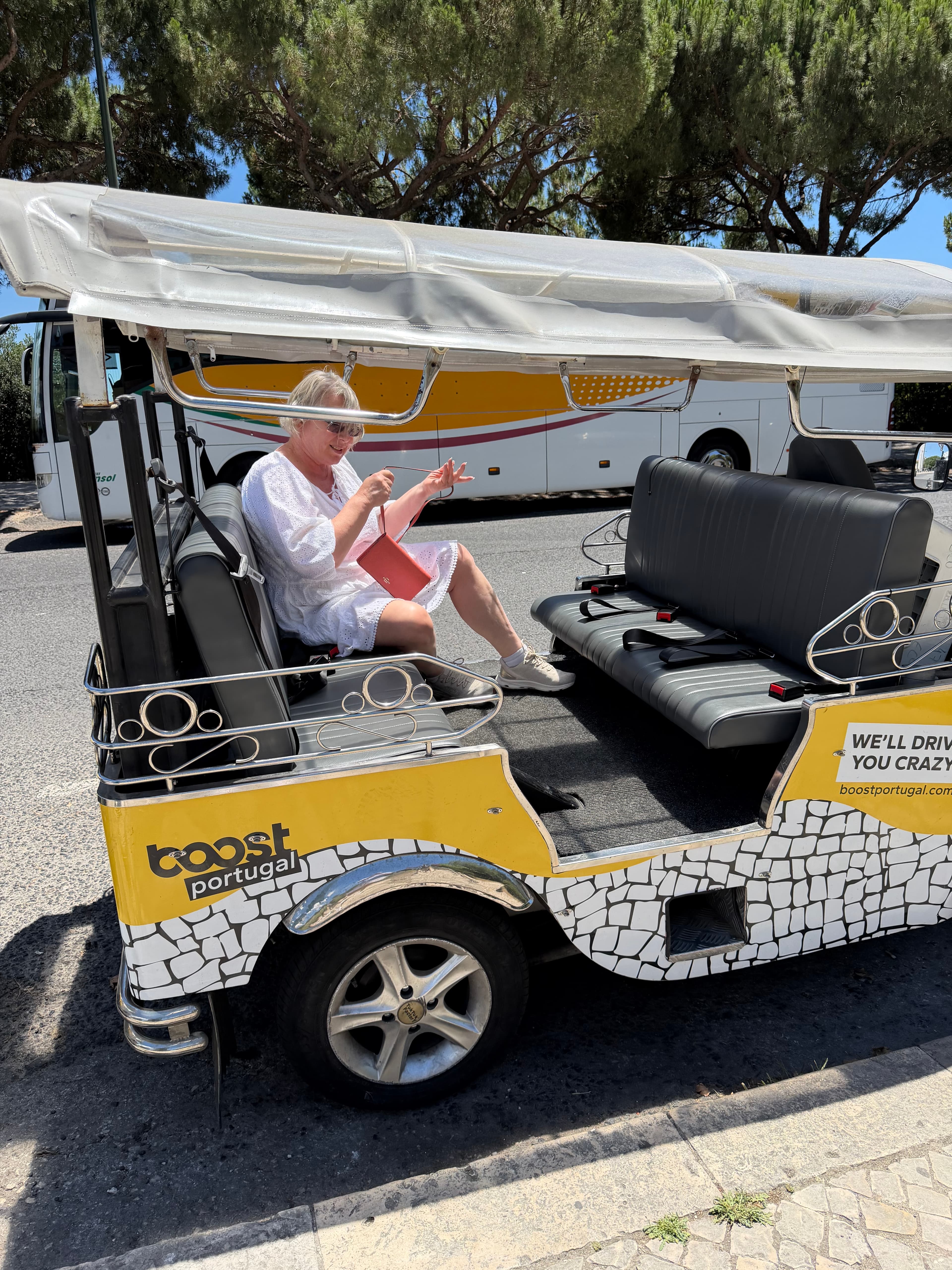 Smiling woman in white dress seated in a yellow-and-white Boost Portugal tuk-tuk tour vehicle on a sunny day, pine trees in b