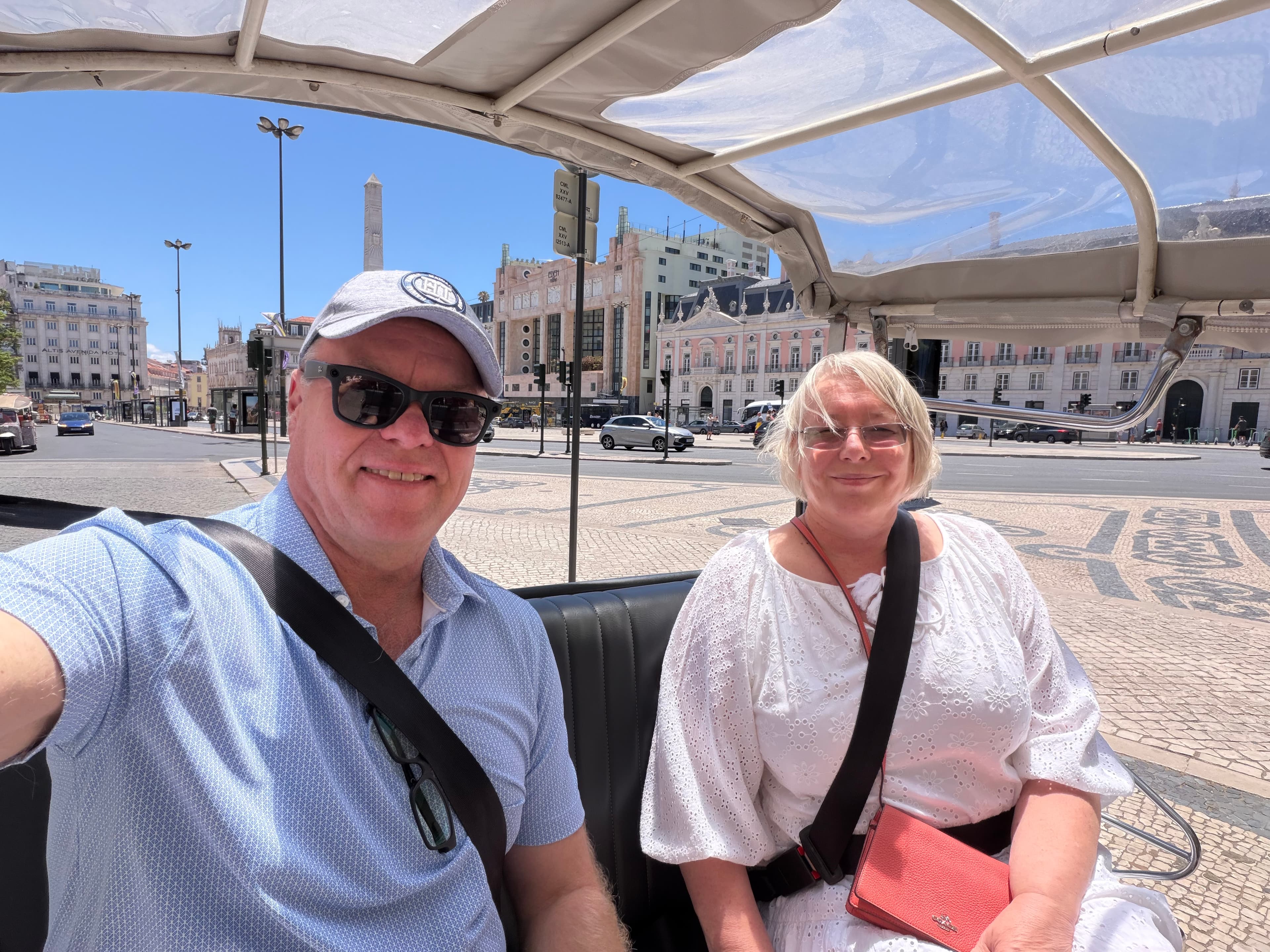 Couple smiling on a tuk-tuk tour in Lisbon's Rossio Square on a sunny day, with ornate pink buildings and an obelisk behind t